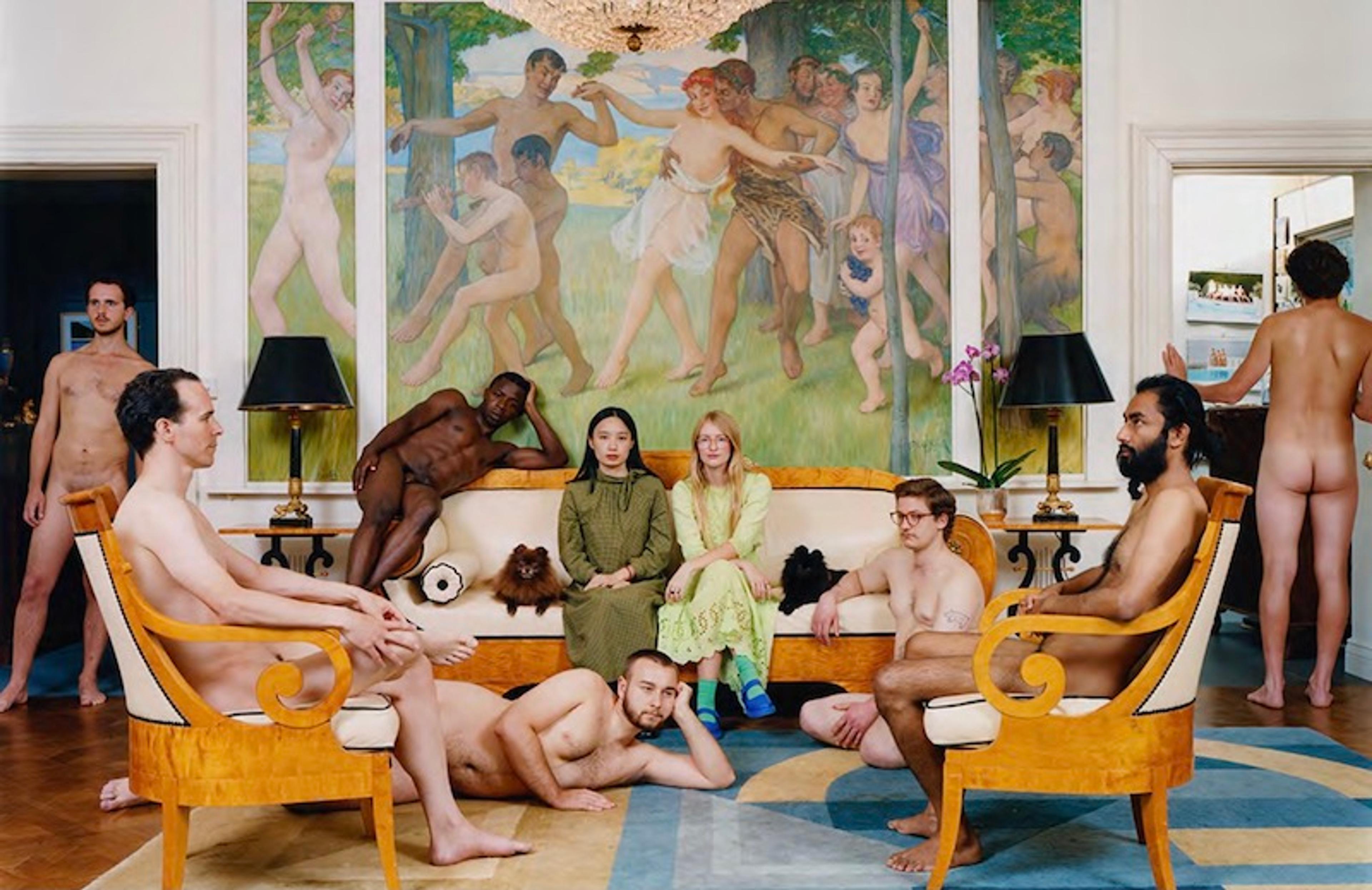 An image of two women wearing green dresses in a living room, surrounded by several nude men