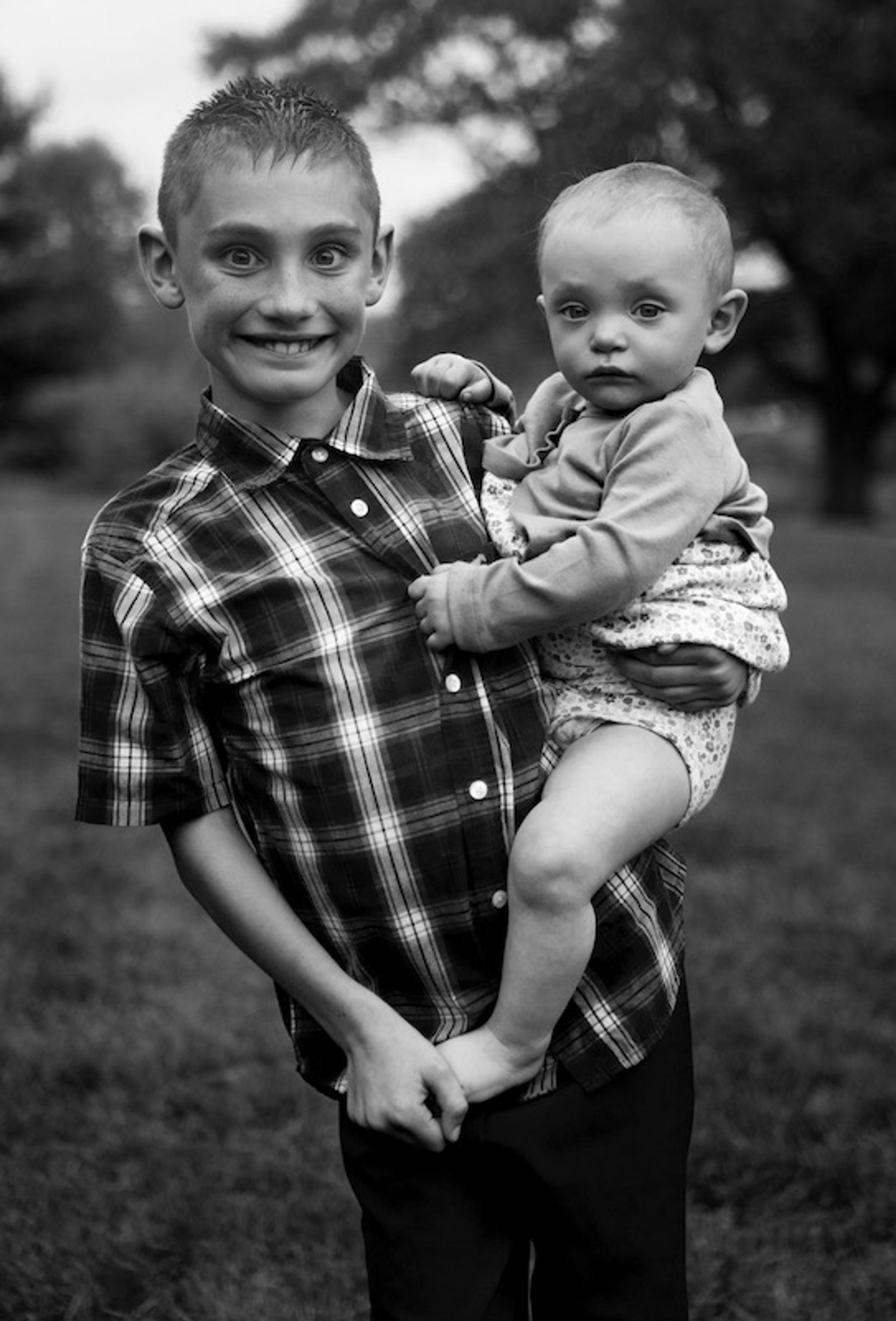 A black and white photograph of a young boy in a plaid shirt smiles and holds a baby on his hip