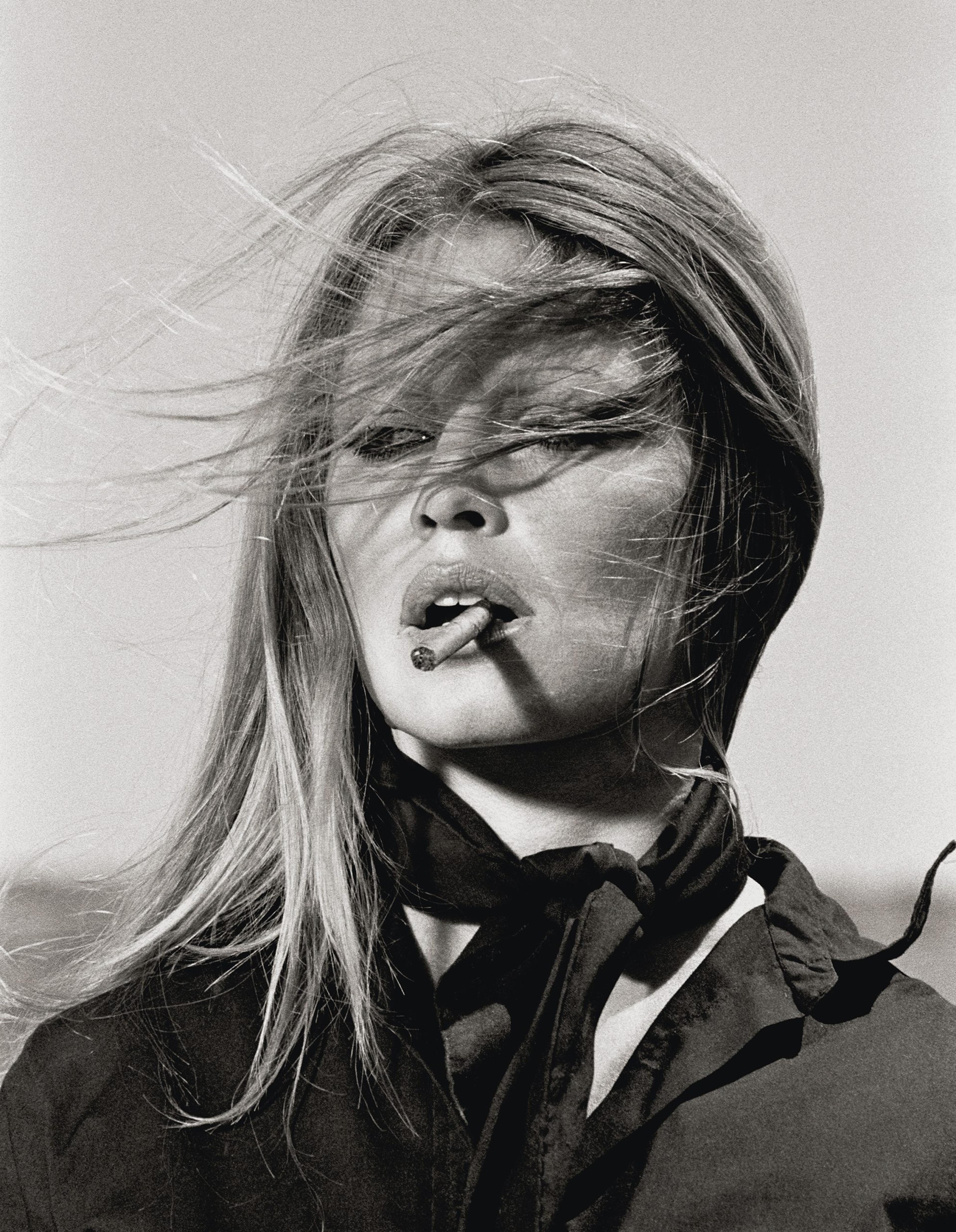 A black-and-white photograph of Brigitte Bardot with windblown hair, wearing dark clothing and holding a cigarette in her mouth