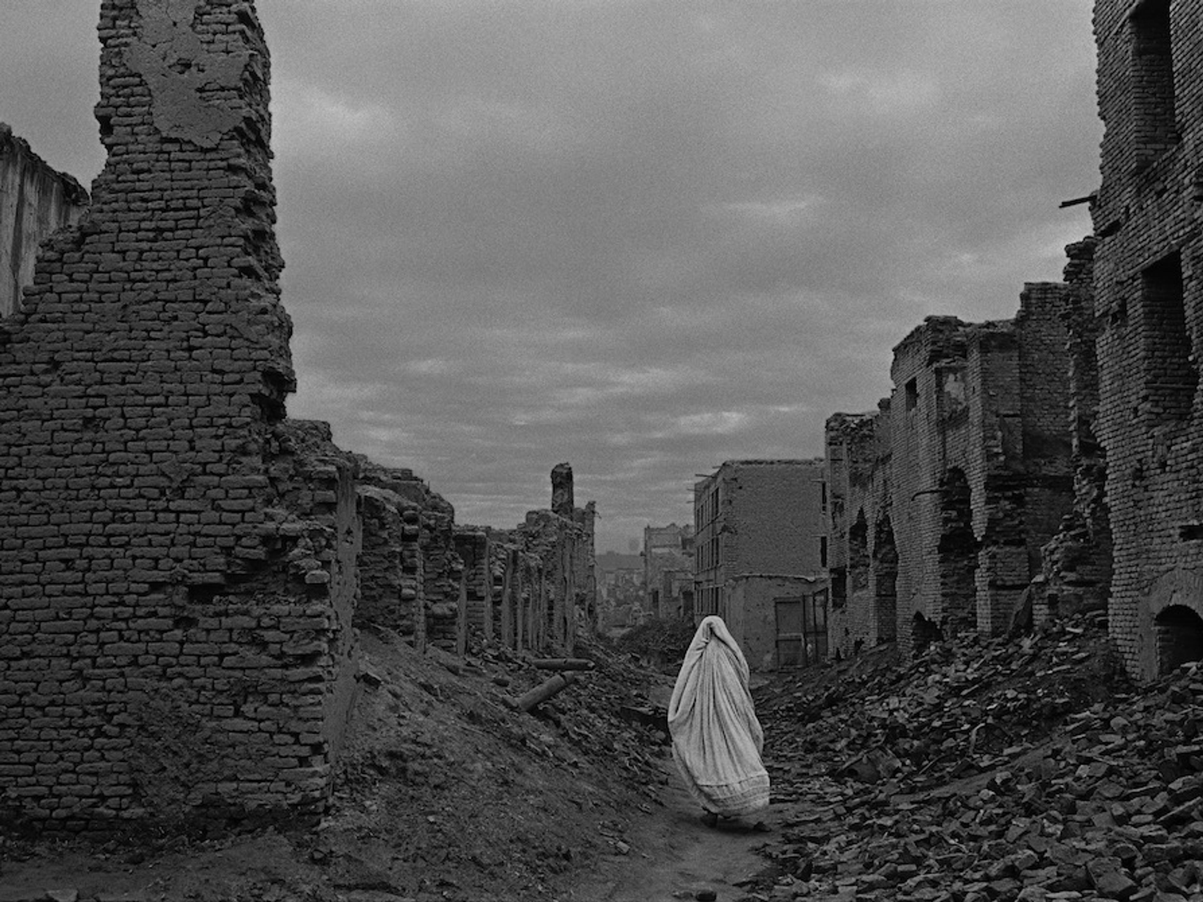 A black-and-white photograph of a figure walking through a desolate landscape, wearing a white, flowing robe