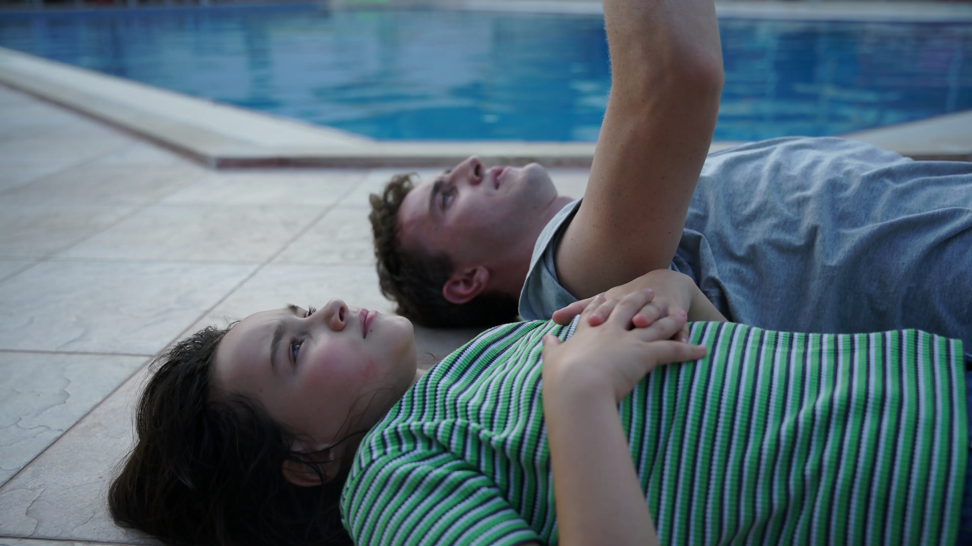 A man and a girl lie on their backs on the ground beside a pool, their heads close together and their gazes directed upwards. The girl holds her hand to her chest. The pool’s blue water is visible in the background.