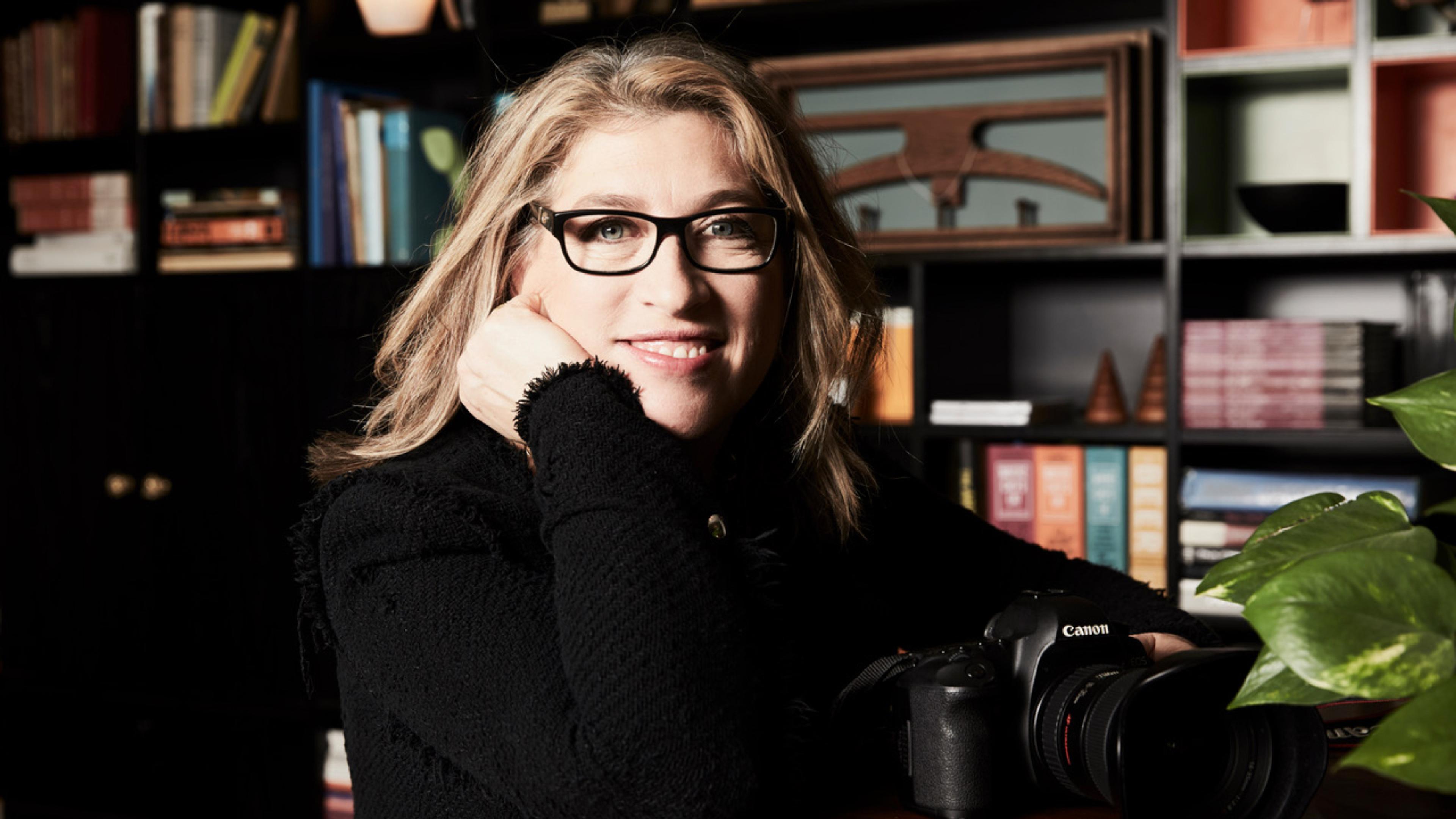 Portrait of photographer Lauren Greenfield in glasses, resting her chin on her hand, with a Canon camera in front of her and bookshelves behind