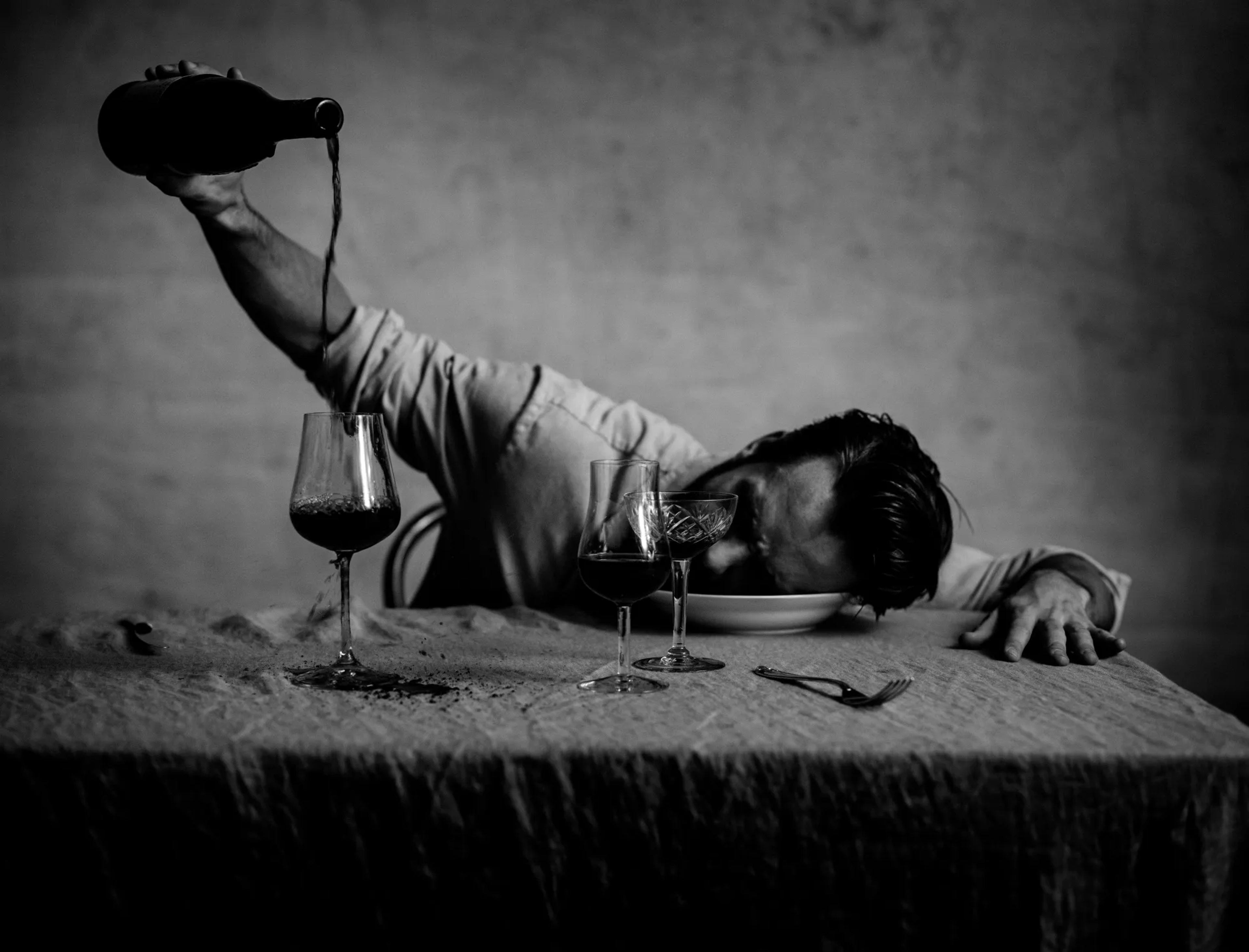 A man sits at a table, pouring wine into glasses while resting his head in a bowl.