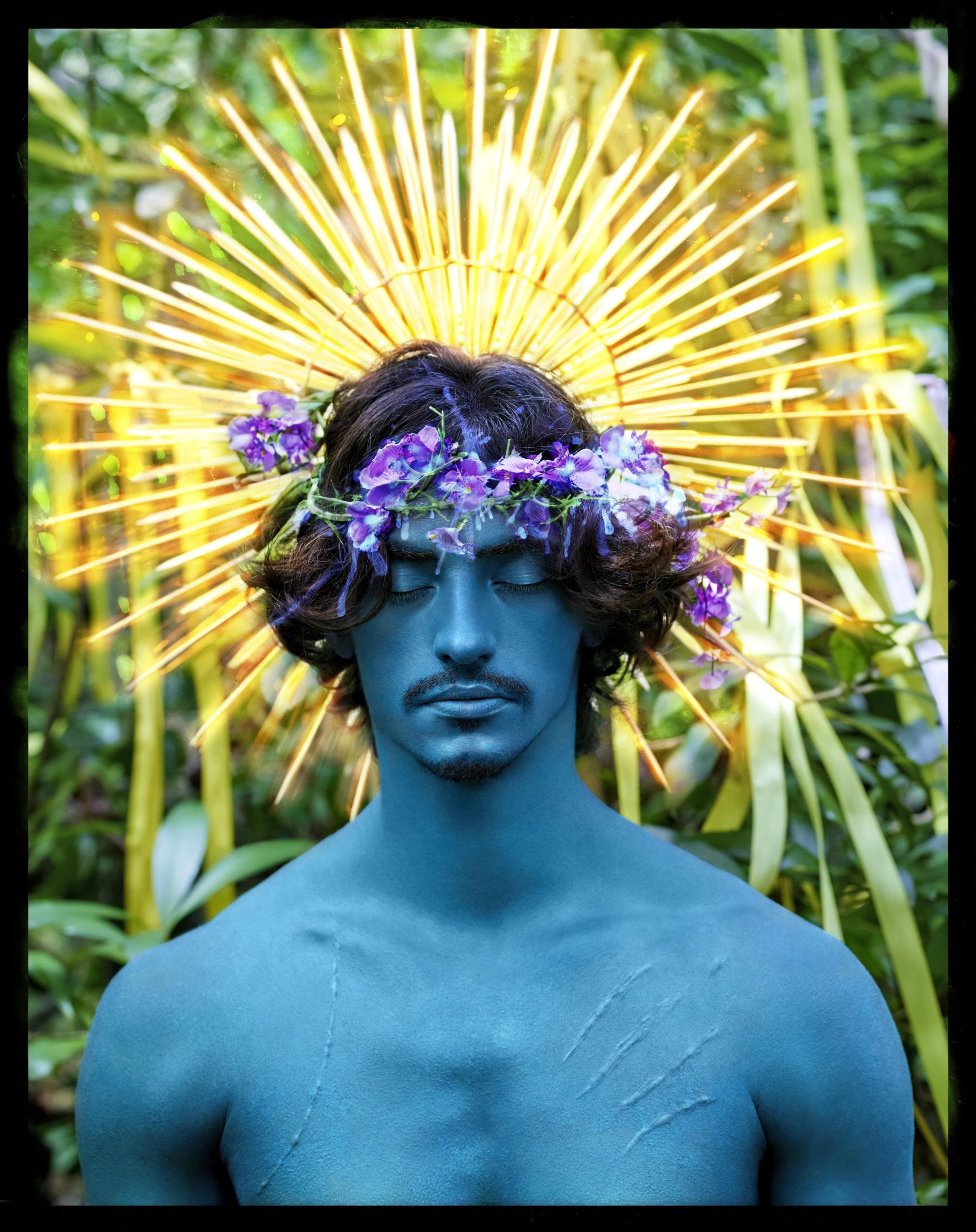 A photograph of a man covered in blue paint, wearing a purple flower crown that exudes light beams