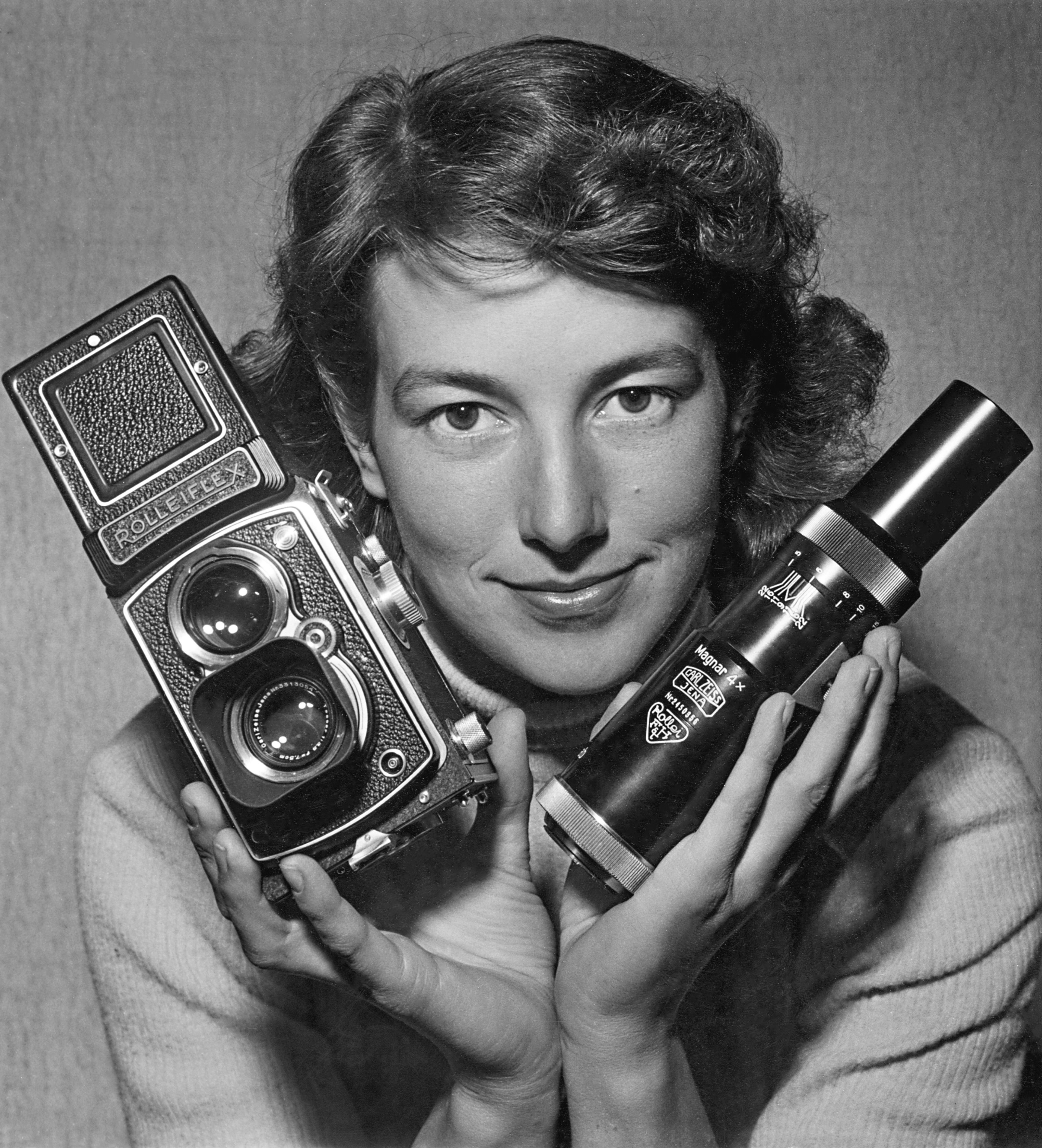 Ett svartvitt porträtt av en leende kvinna som håller upp en klassisk Rolleiflex tvåögd spegelreflexkamera i sin högra hand och ett stort kameraobjektiv (Carl Zeiss Magnar 4x) i sin vänstra hand.
