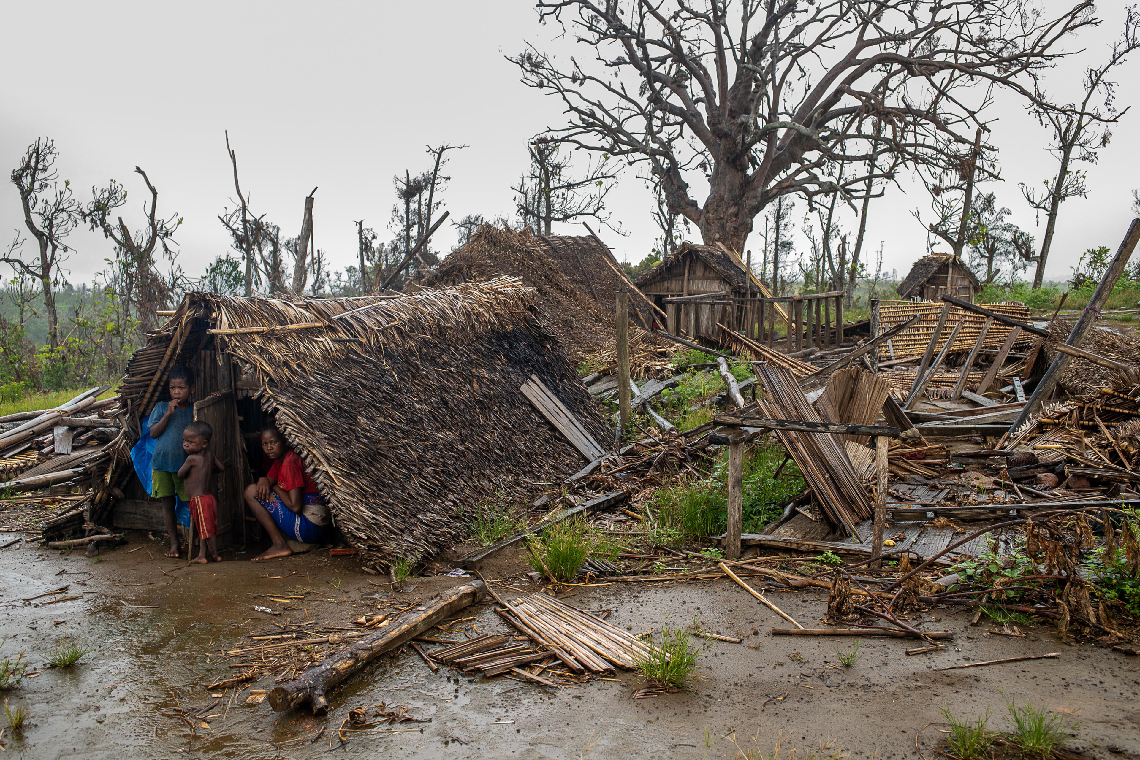 Three children under a straw hut seeking cover from rain. Muddy ground in the foreground and destructed huts to the right. Trees and cloudy skies in the background.
