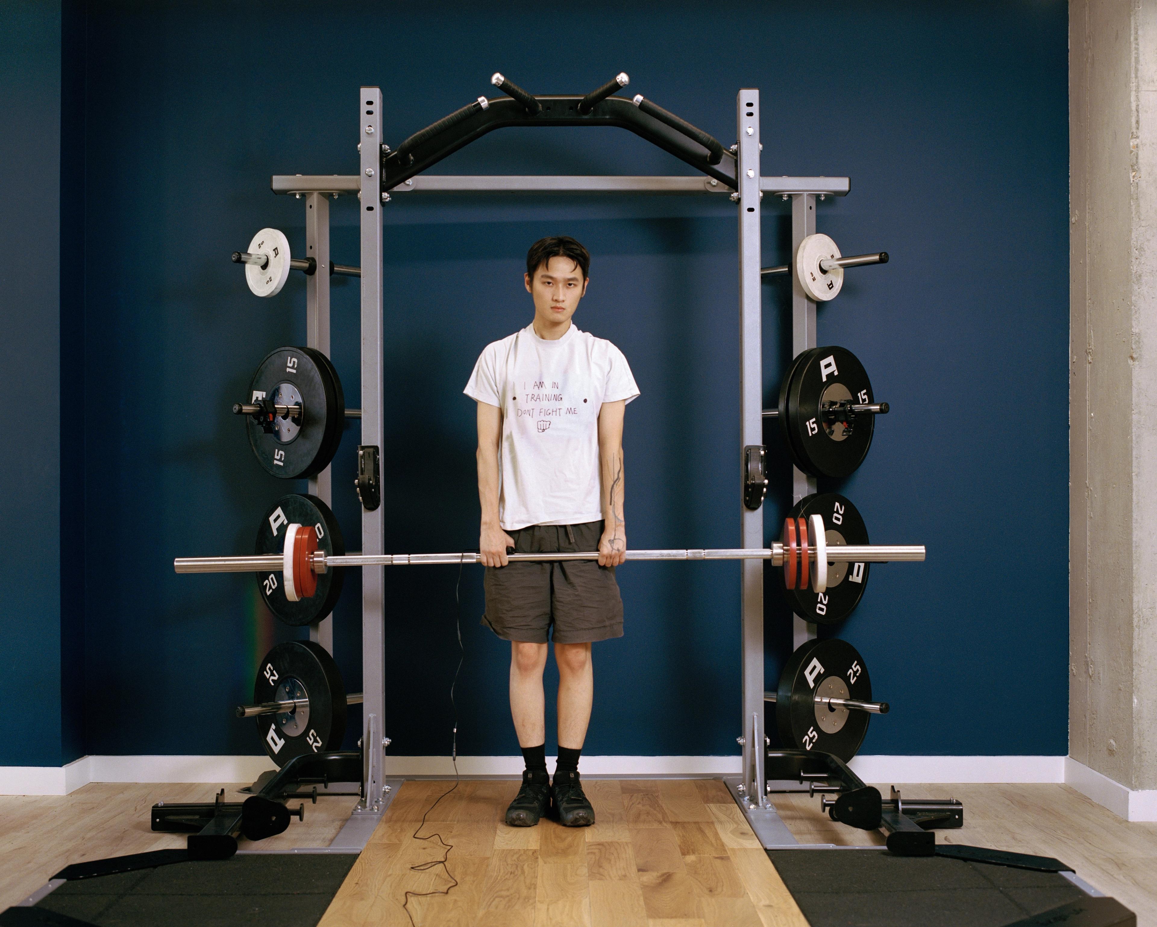  En stående bild som visar en ung man med östasiatiskt utseende och korta, mörka frisyr, centrerad i ett squat-rack av metall i ett gym. Han har en vit t-shirt med trycket "I AM IN TRAINING DON'T FIGHT ME" och svarta shorts. Han håller en skivstång som vilar på korta säkerhetsarmar, med flera viktskivor på varje sida. Bakom honom finns en mörkblå vägg och ett golv av ljust trä. Han tittar direkt mot kameran med ett allvarligt uttryck.