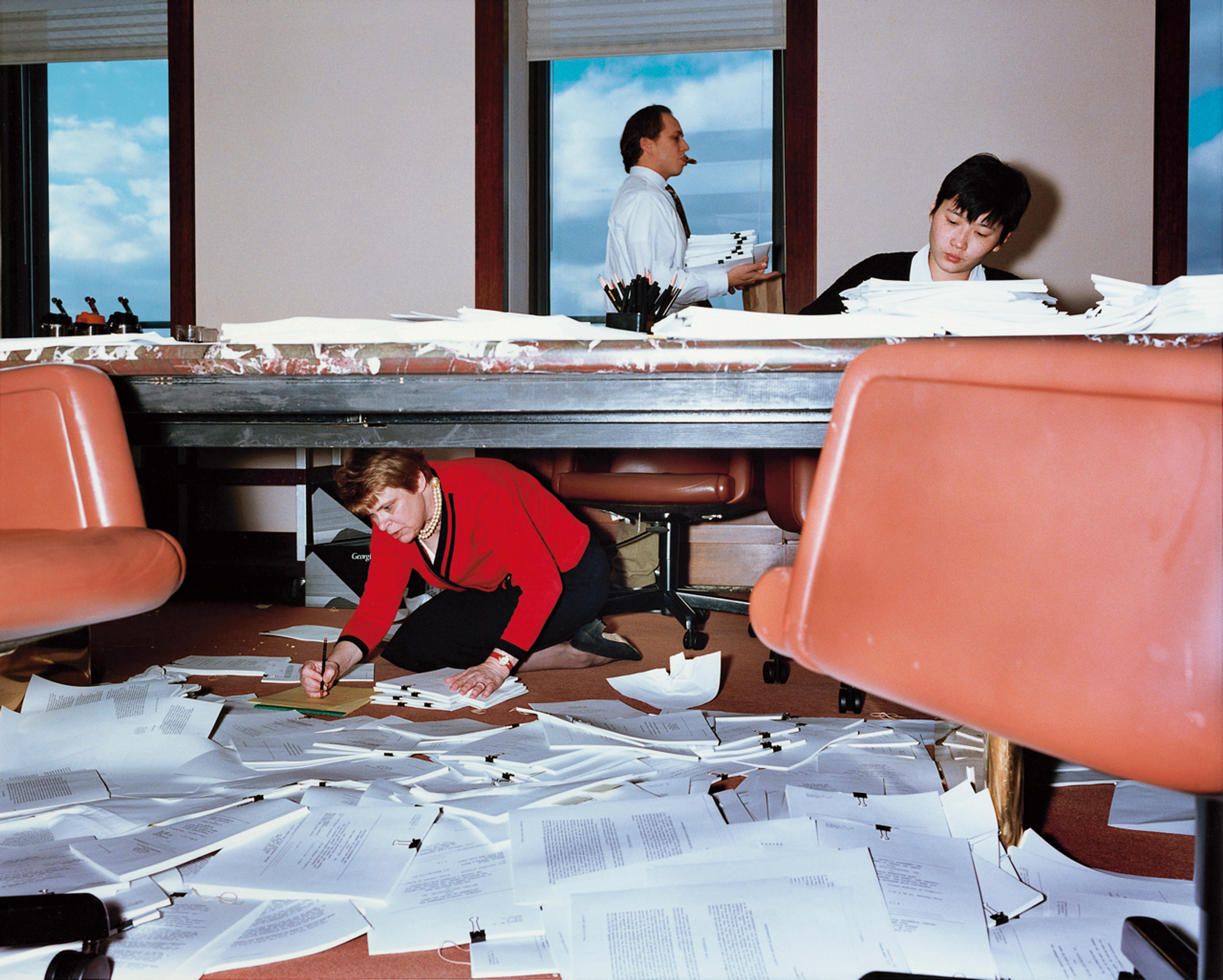 Office scene with papers scattered across the floor, one person working under the table while two colleagues work above.