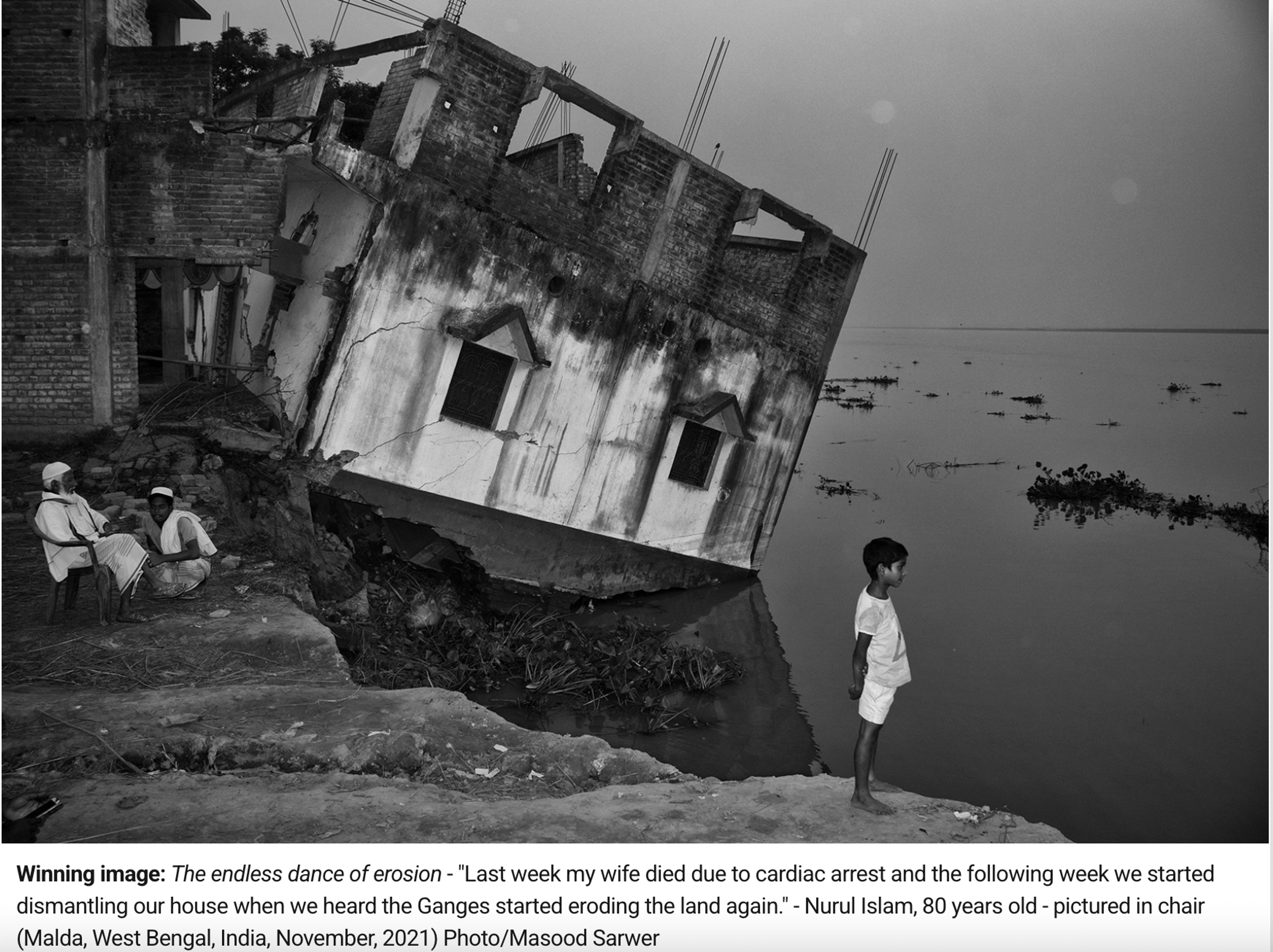 Black and white image of a deteriorated two stories high building on the shores of river Ganges, slowly collapsing into the river. Young boy in white clothing standing by the shoreline in the middle-right and two elderly men in white clothing sitting next to each other to the far left. At the bottom of the image there's a caption saying: "Winning image: The endless dance of erosion - "Last week my wife died due to cardiac arrest and the following week we stared dismantling our house when we heard the Ganges started eroding the land again." - Nurul Islam, 80 years old - pictures in chair. "Malda, West Bengal, India, November, 2021) Photo/Masood Sarwer"