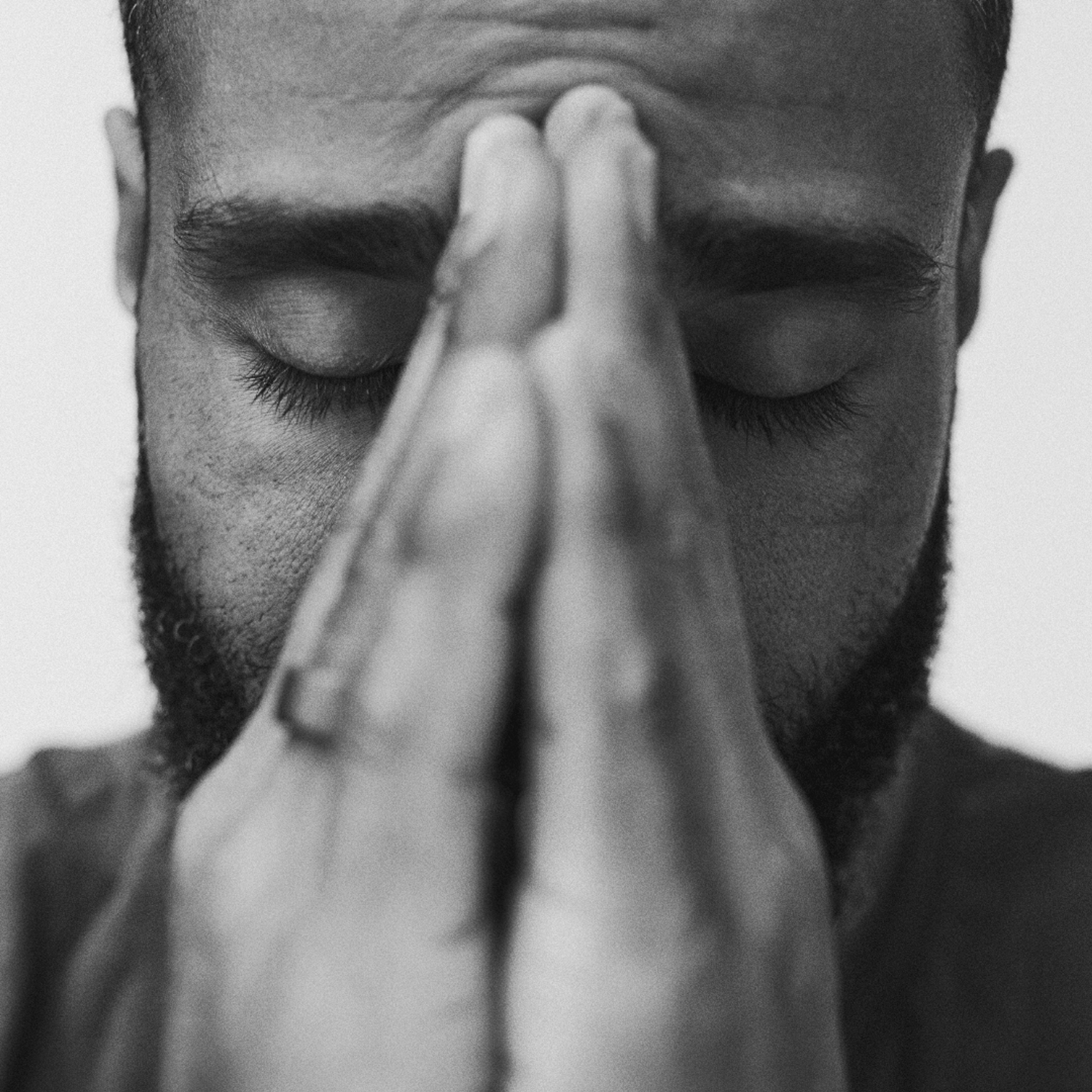 A black-and-white close-up portrait of a man with a beard and short hair. His eyes are closed and his hands are pressed together in front of his face, with his fingertips resting against his forehead in deep focus, prayer, or contemplation.