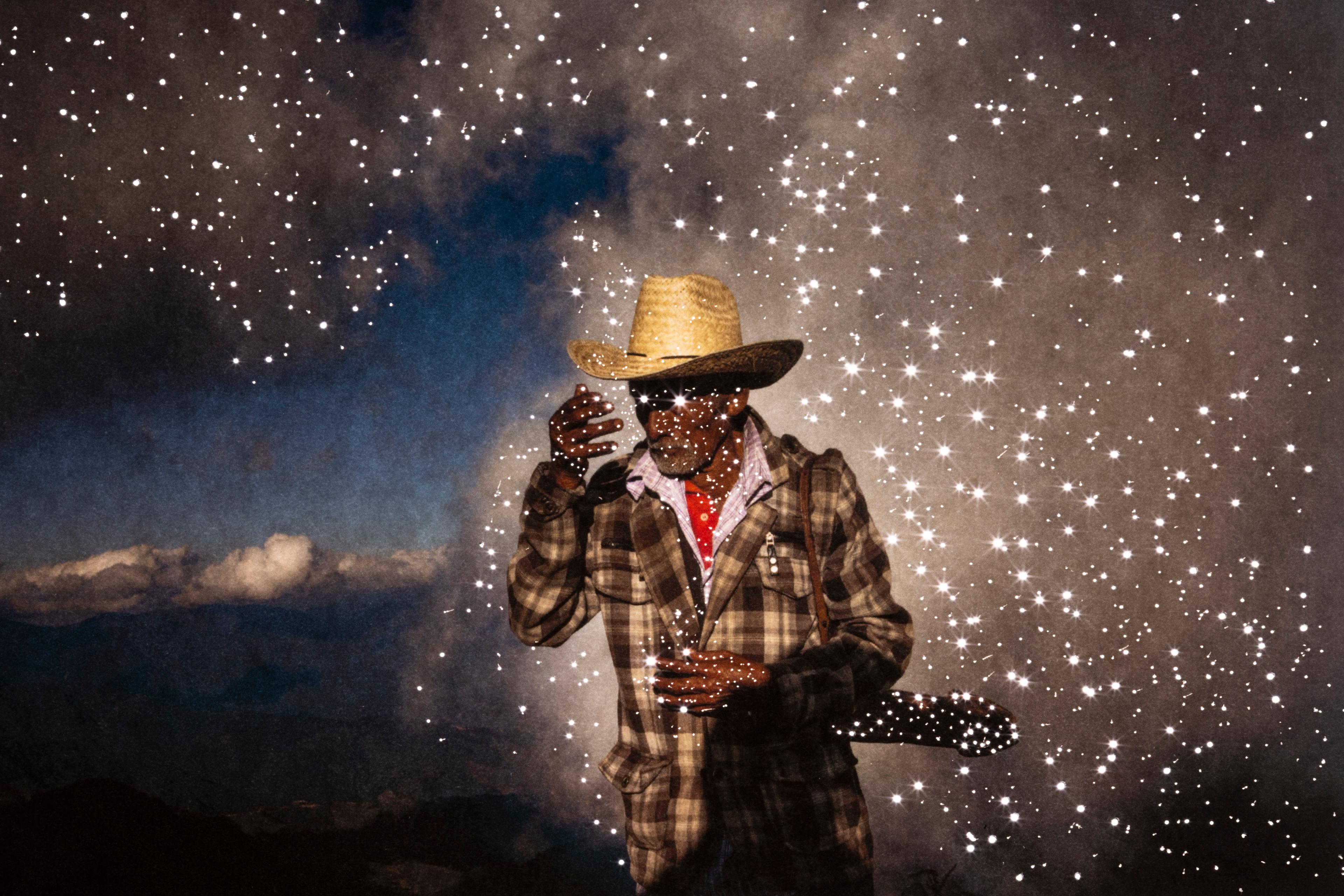 En man i cowboyhatt och kostym som poserar. Han står bakom en stjärnklar himmel och har stjärnor runtomkring sig..