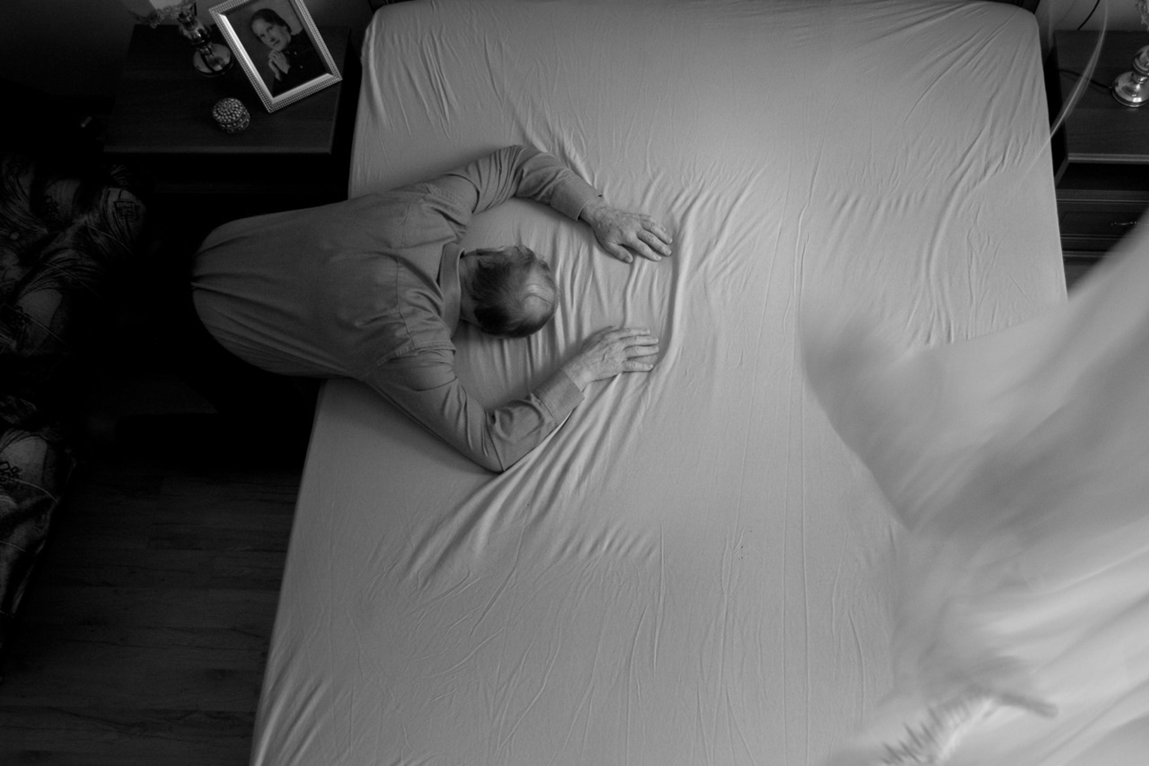 Black-and-white overhead shot of an elderly person curled up on an unmade bed, head bowed with hands pressed against the sheet. A blurred white figure moves through the foreground. A framed photo sits on the nightstand.