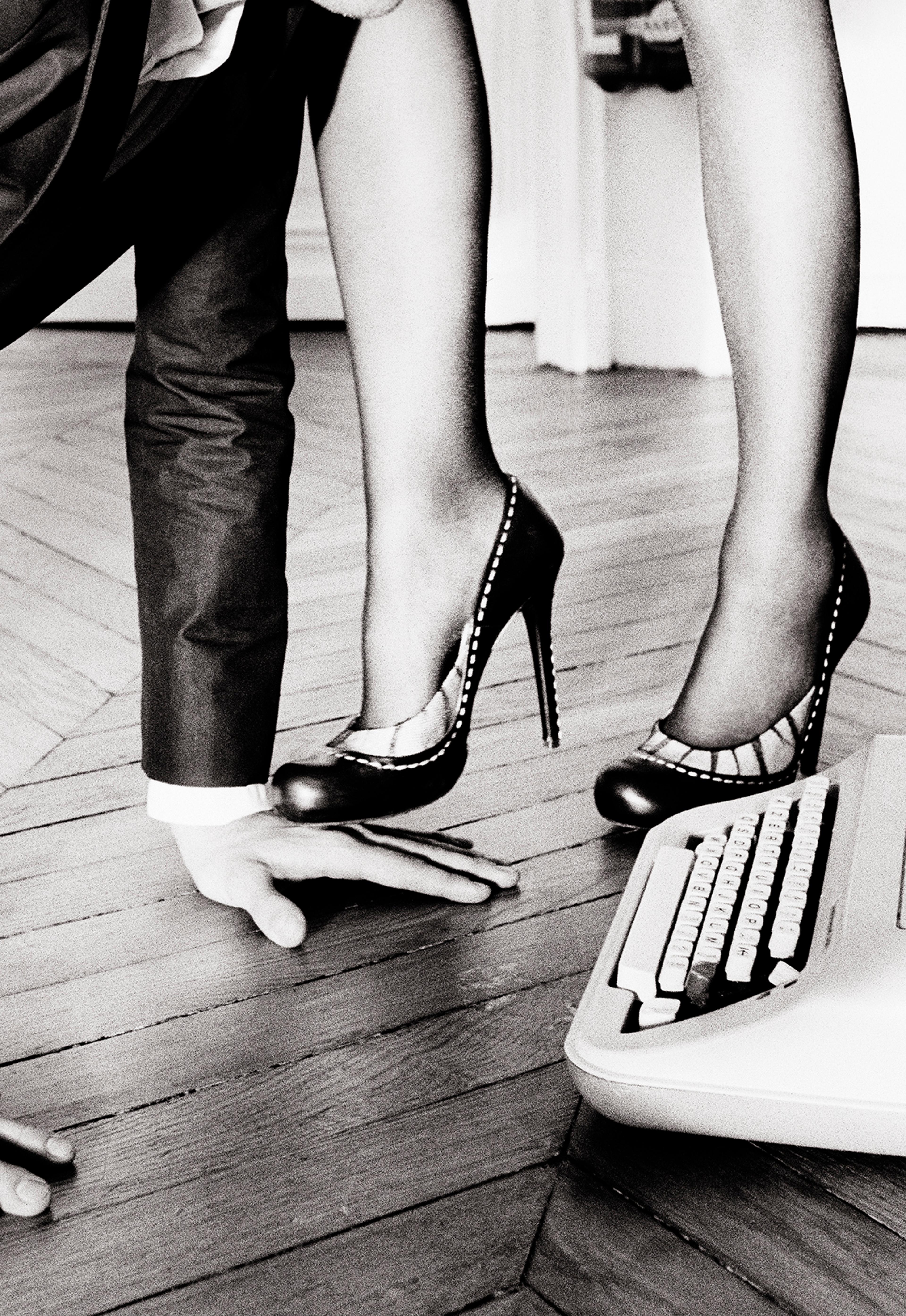 A black and white photograph of a woman's high-heeled toe stepping on a man's hand on the floor
