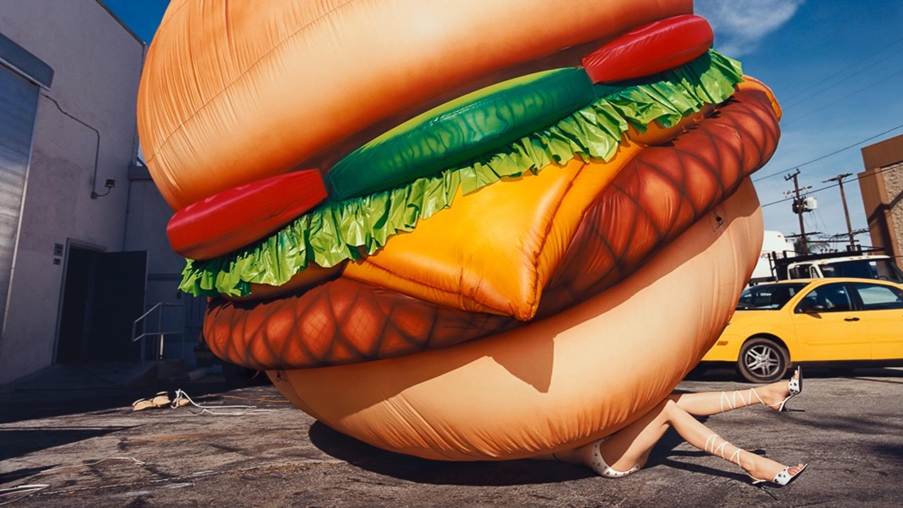 A photo of a woman laying down beneath a large blow-up cheeseburger