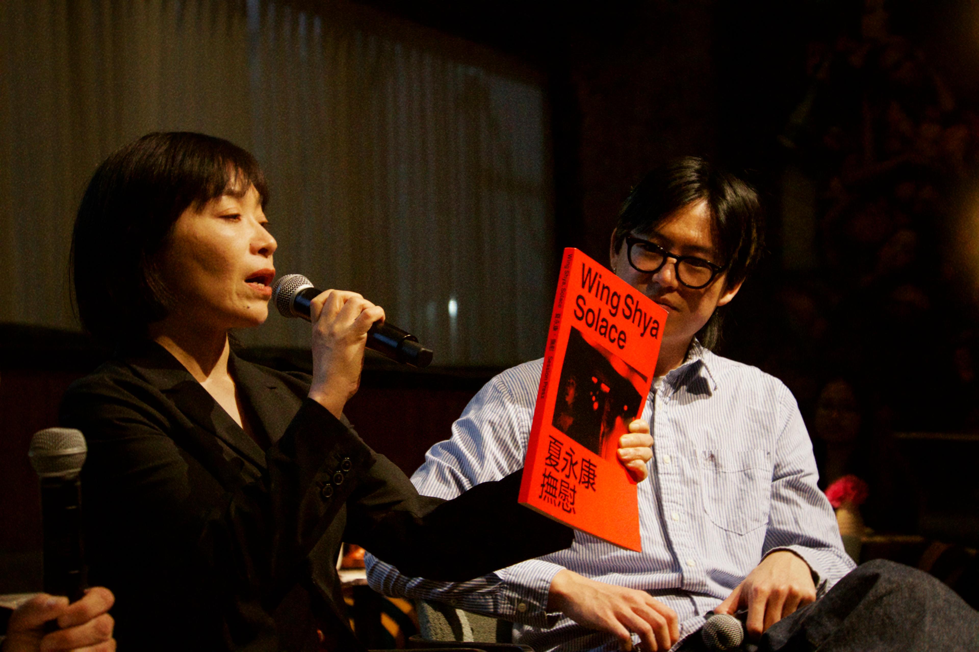 Miwa speaks into a microphone and holds up a red book called "Wing Shya: Solace: