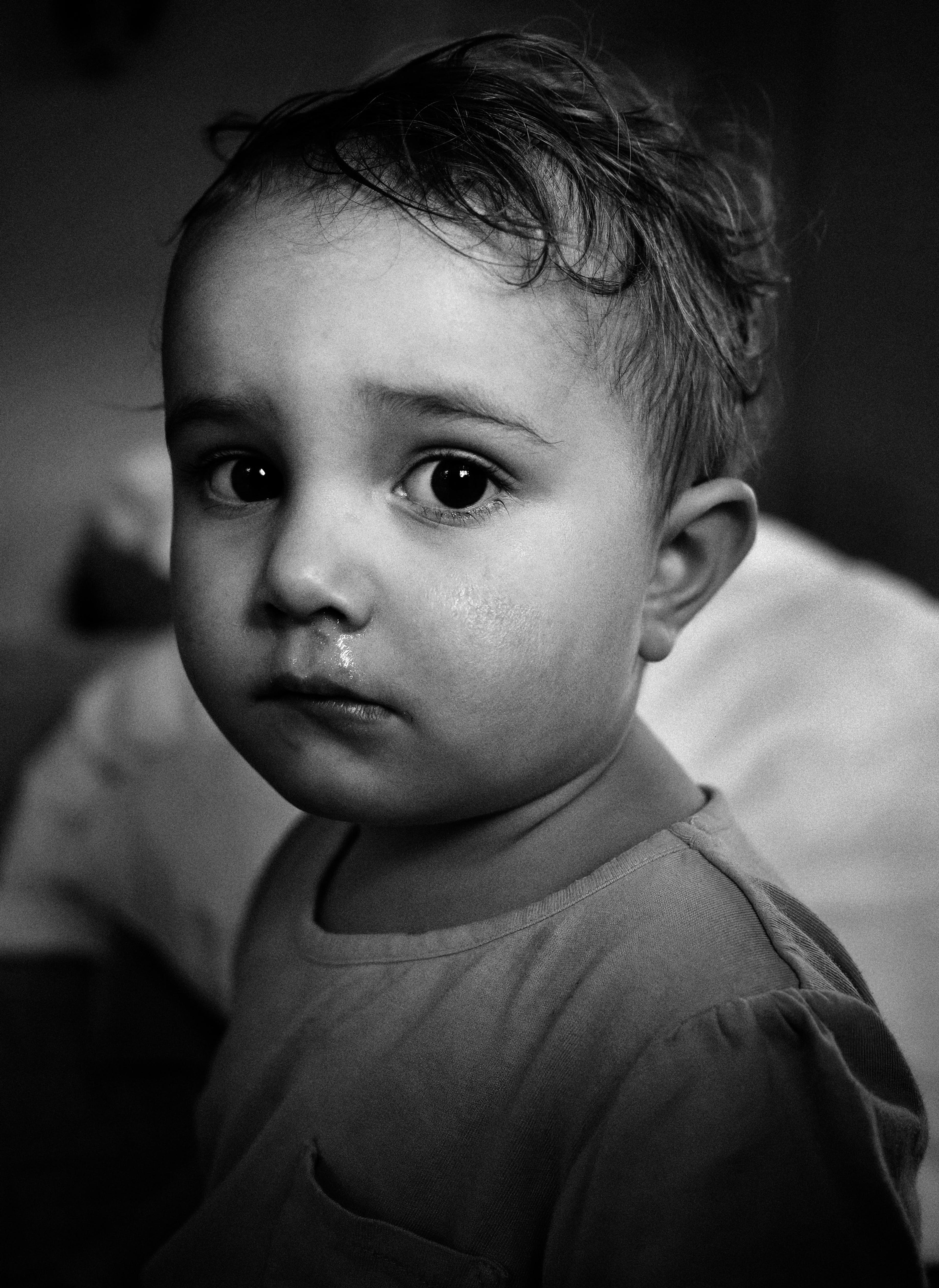 A close-up black and white photograph of a small toddler with large, dark eyes gazing thoughtfully at the camera. The child has a runny nose and a calm, quiet expression.