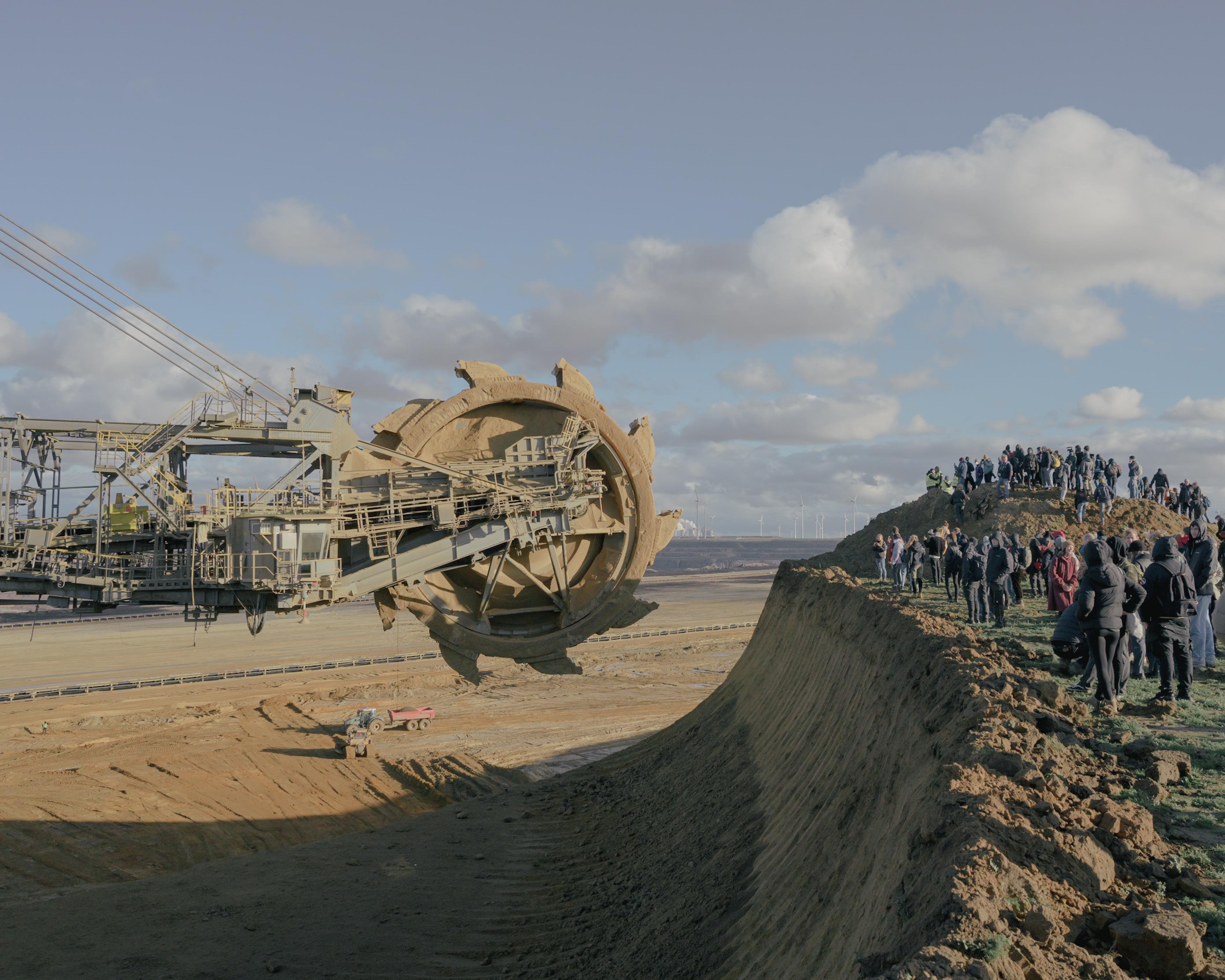 Large drilling apparatus to the left facing a crowded audience standing on the shelf of a crater. Blue and cloudy skies in the background.