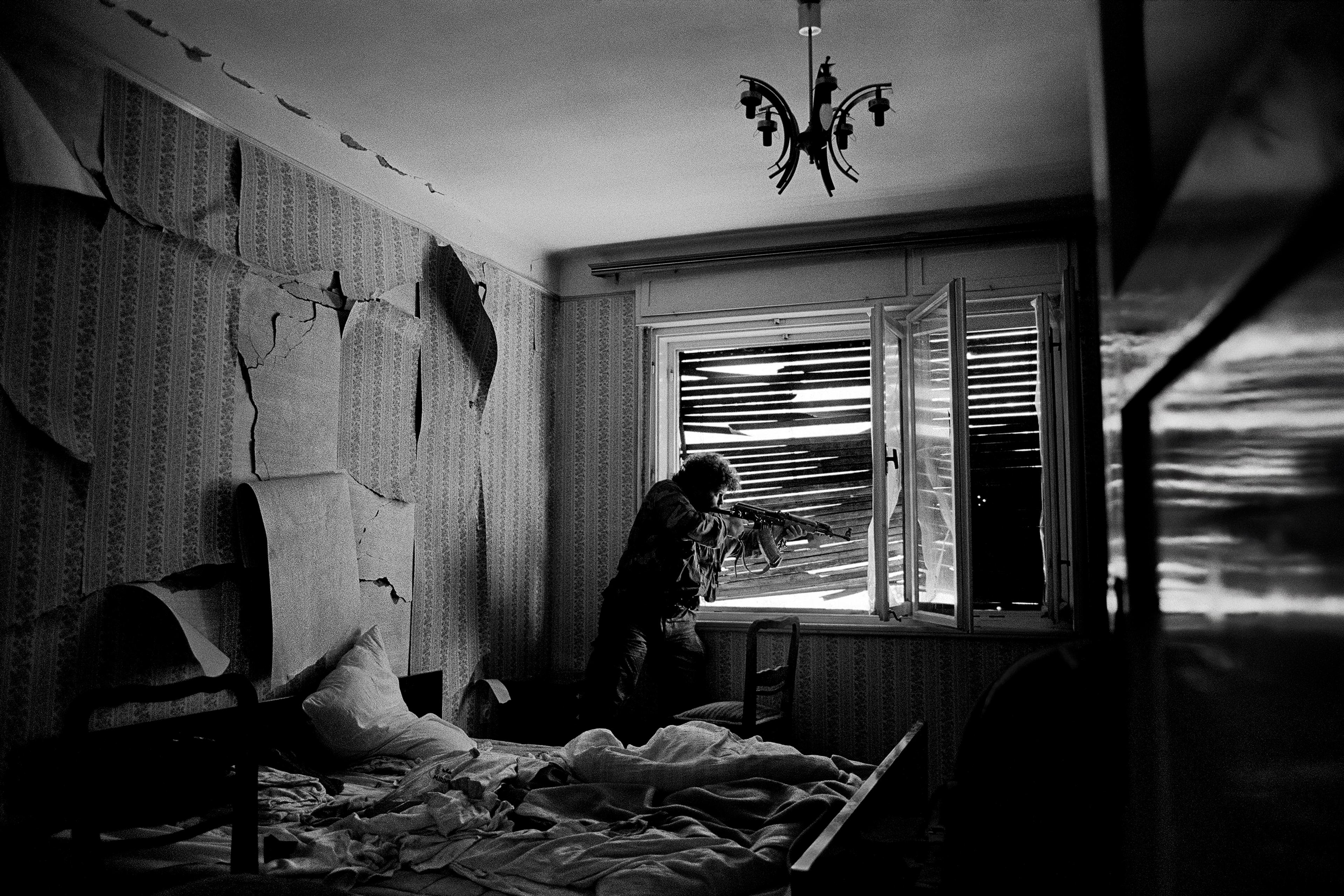 B/W photo of a destroyed room with man standing next to the window holding an AK47