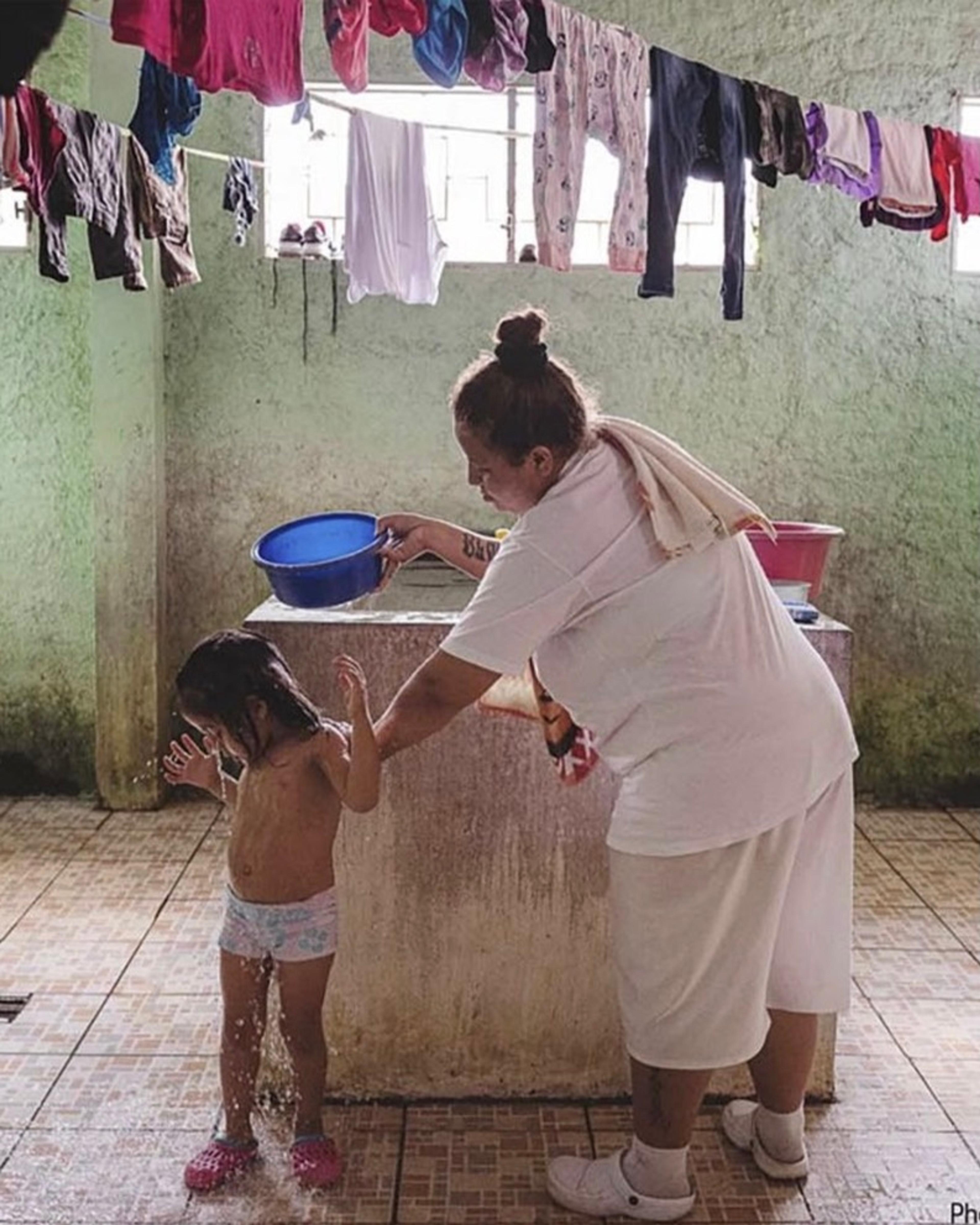 A photograph of a woman bathing a child with a blue bowl, clothes hanging in the background