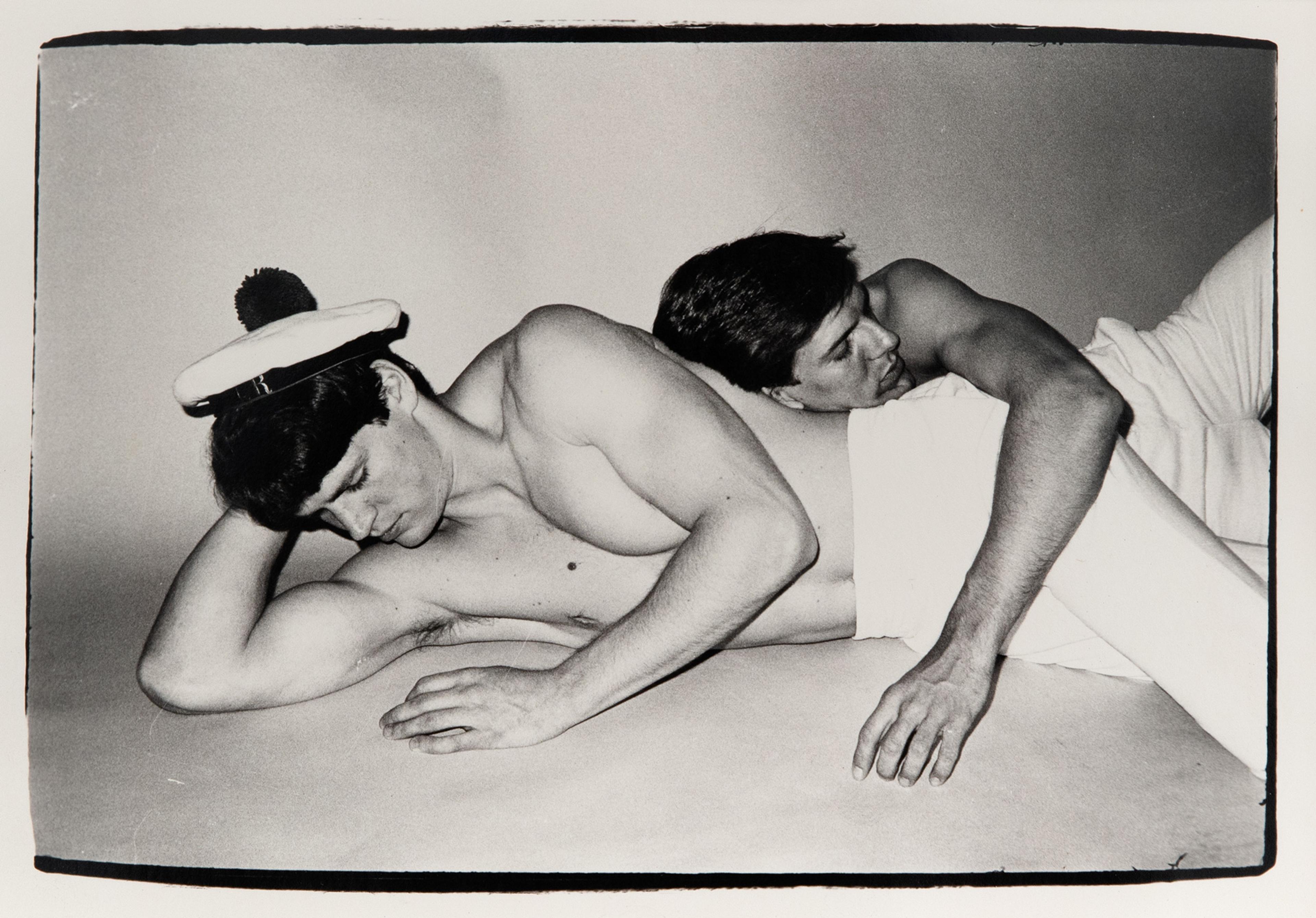 One topless man with a sailor hat lying on the floor, another topless man leaning on him with his head.