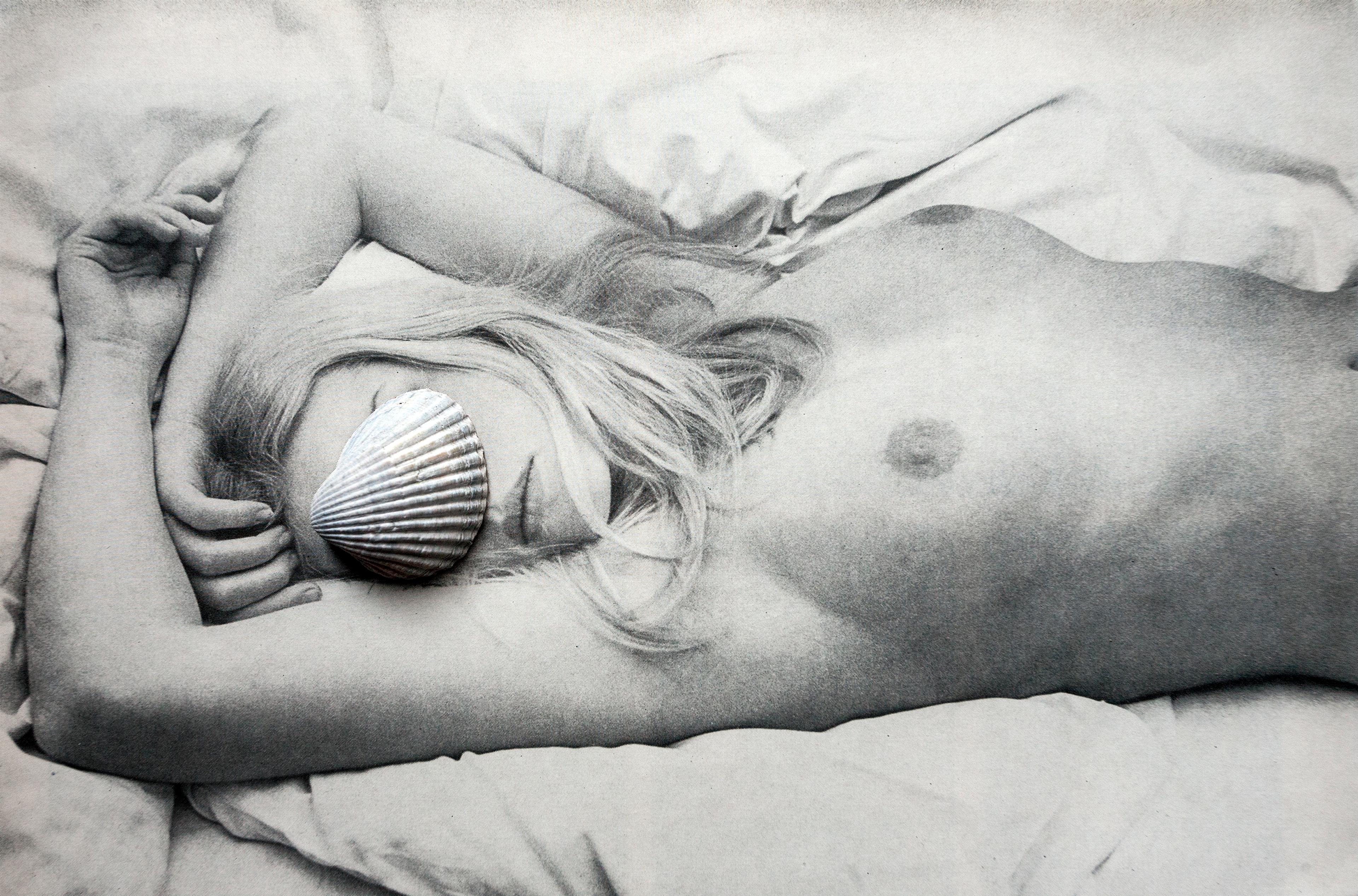  Black-and-white photograph of a nude woman lying on her back on a bed. Her arms are bent above her head, and her blonde hair spreads across the pillow. A seashell covers one of her eyes, creating a surreal and artistic expression.