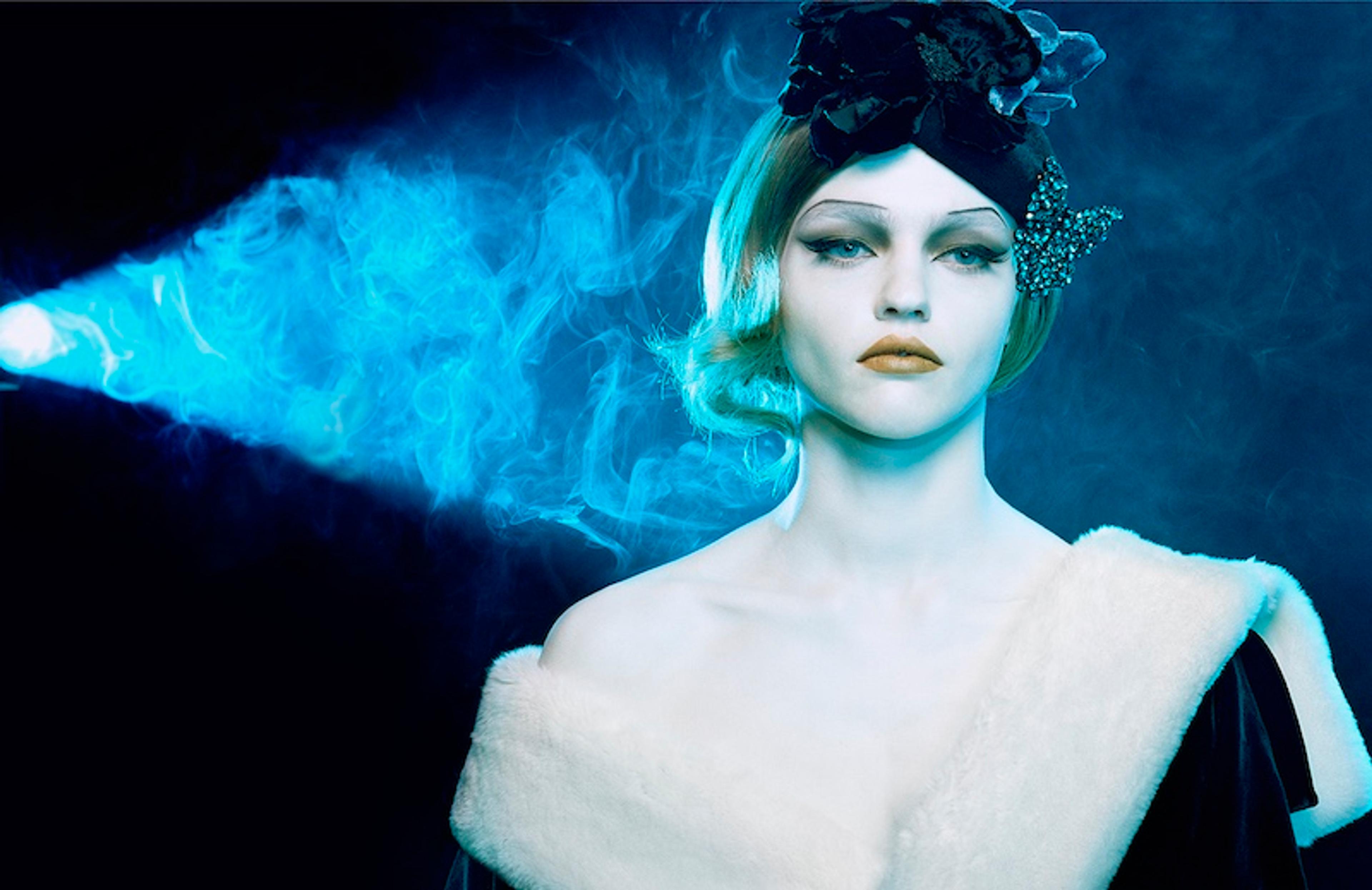 A photograph of a pale woman wearing a fur coat and flapper-style headdress, surrounded by smoke and illuminated by a blue light