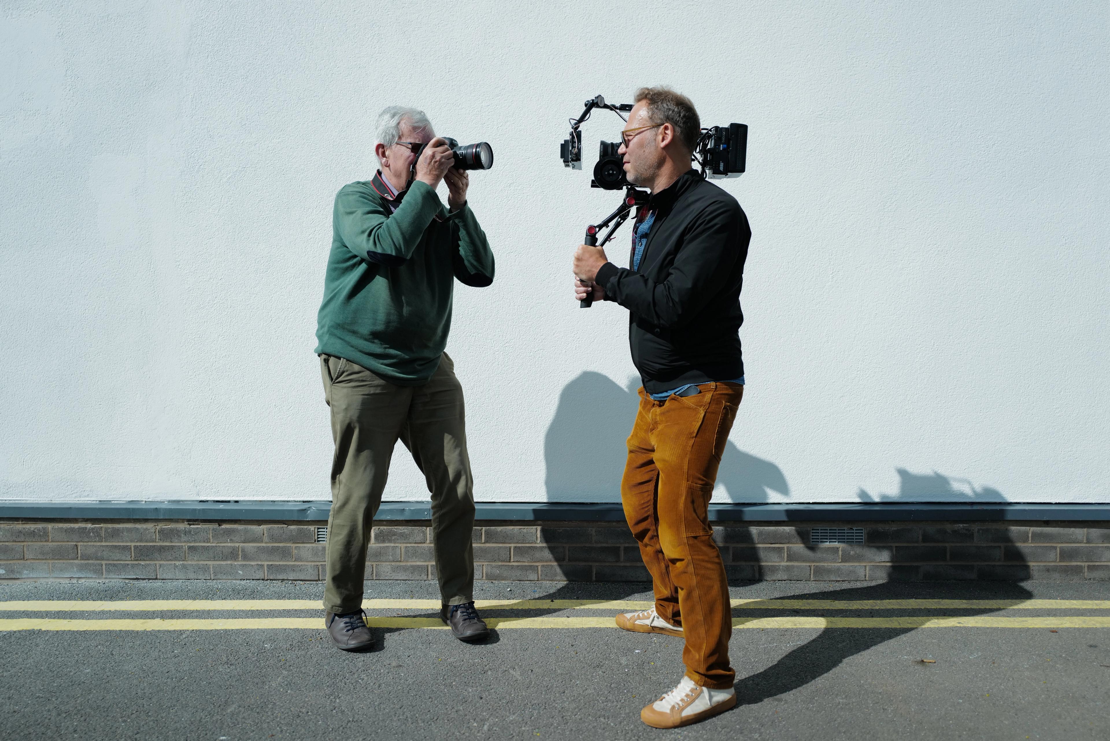 Two men photographing and filming each other outdoors in front of a white wall. An older man on the left holds a DSLR camera, while a younger man on the right holds a video camera on a rig.