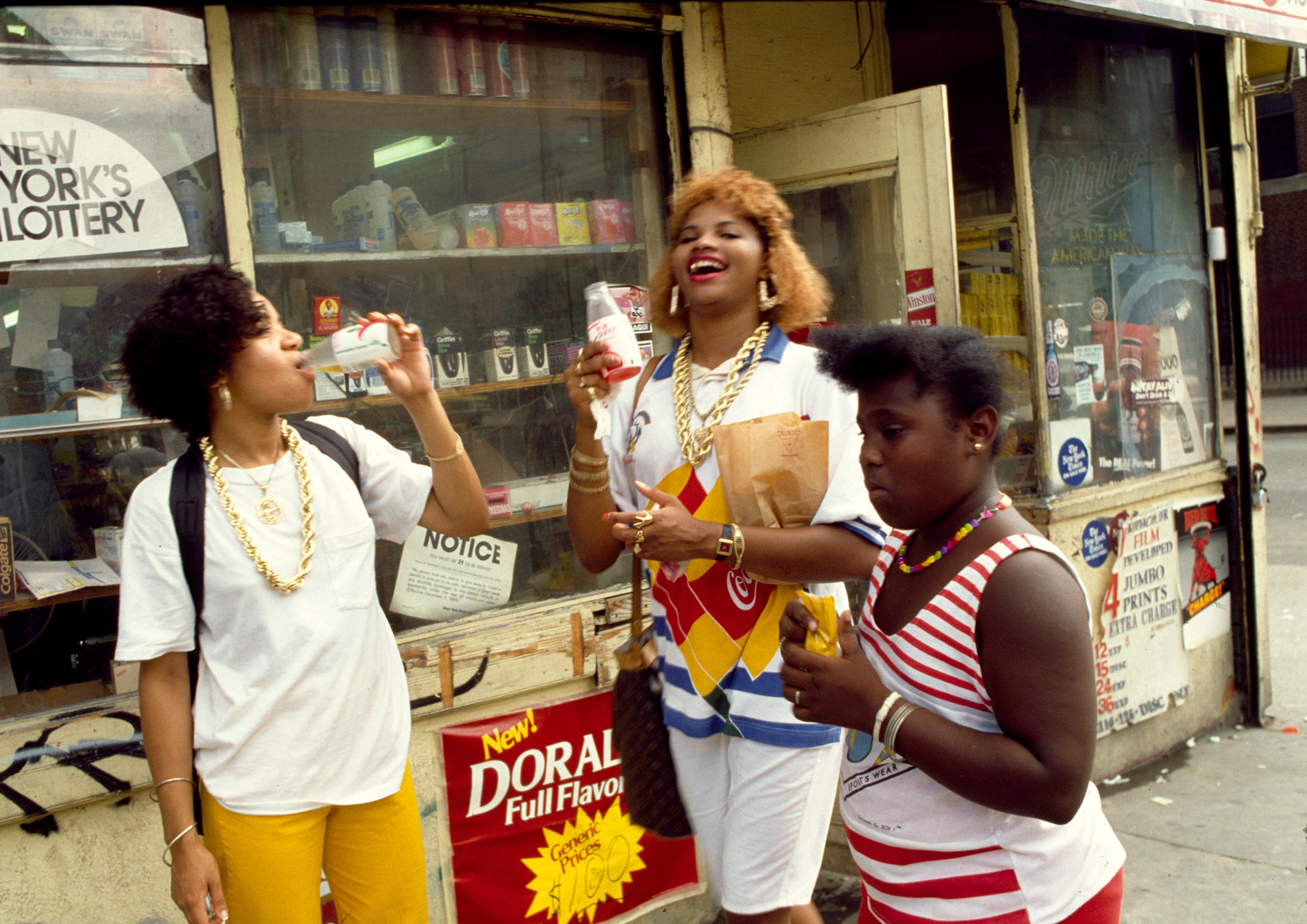 Three women stand outside of a bodega drinking sodas and eating snacks