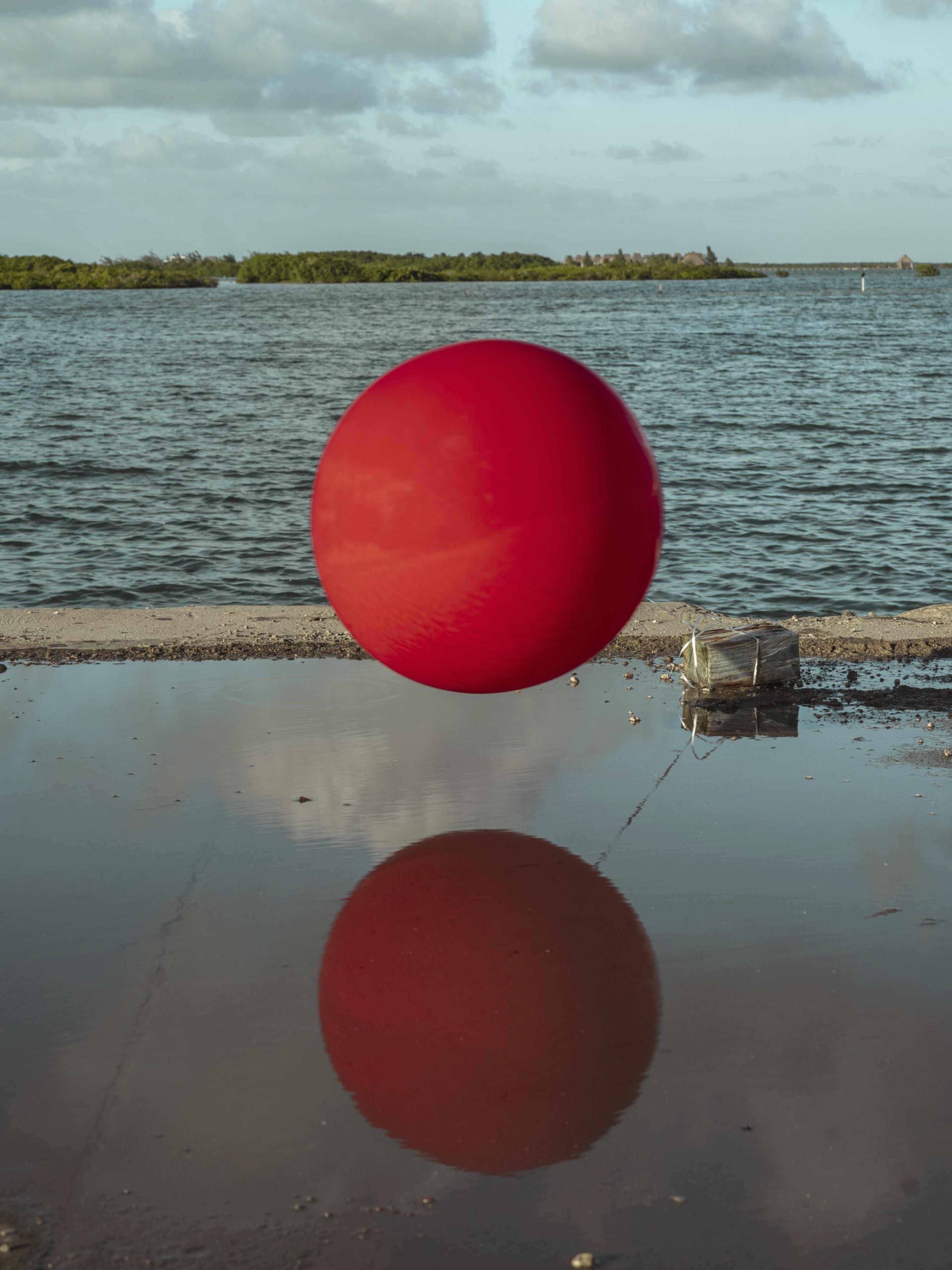A photograph of a red sphere in the foreground with a body of water in the background.