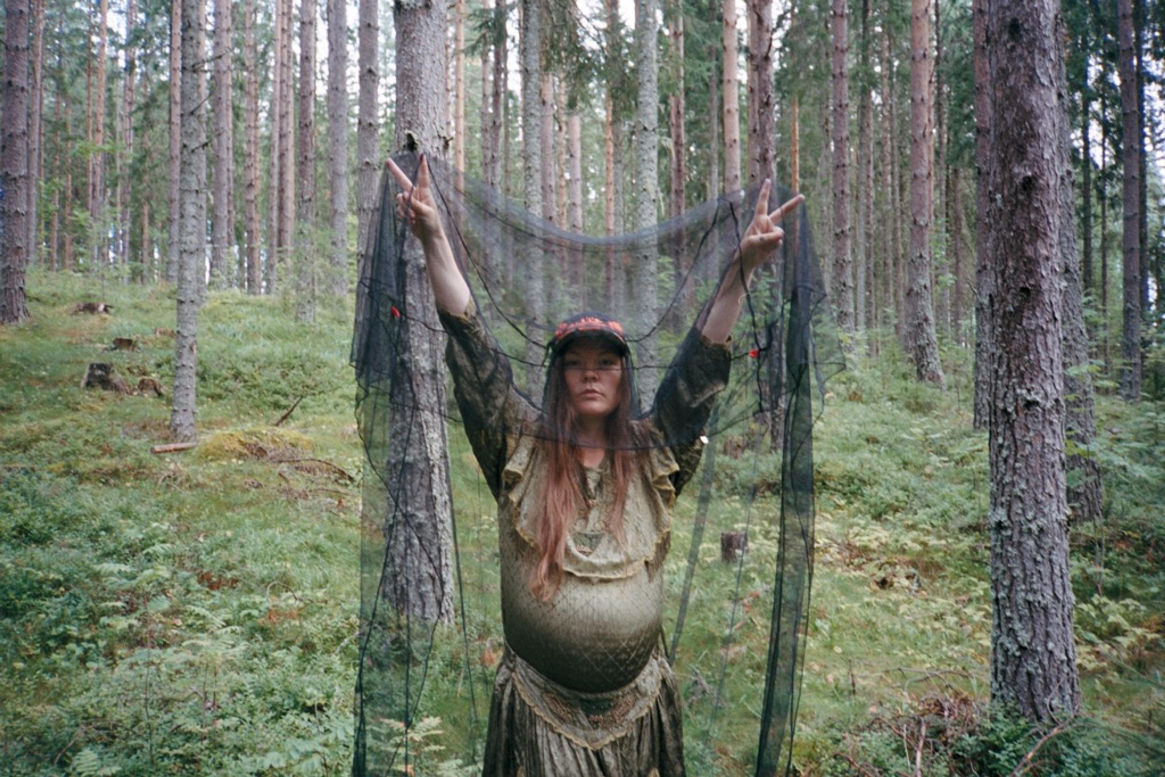 A pregnant woman stands in a forest, holding up a large, black net over herself with both hands while making peace signs. She is wearing a greenish-yellow sparkling dress and a baseball cap on her head.