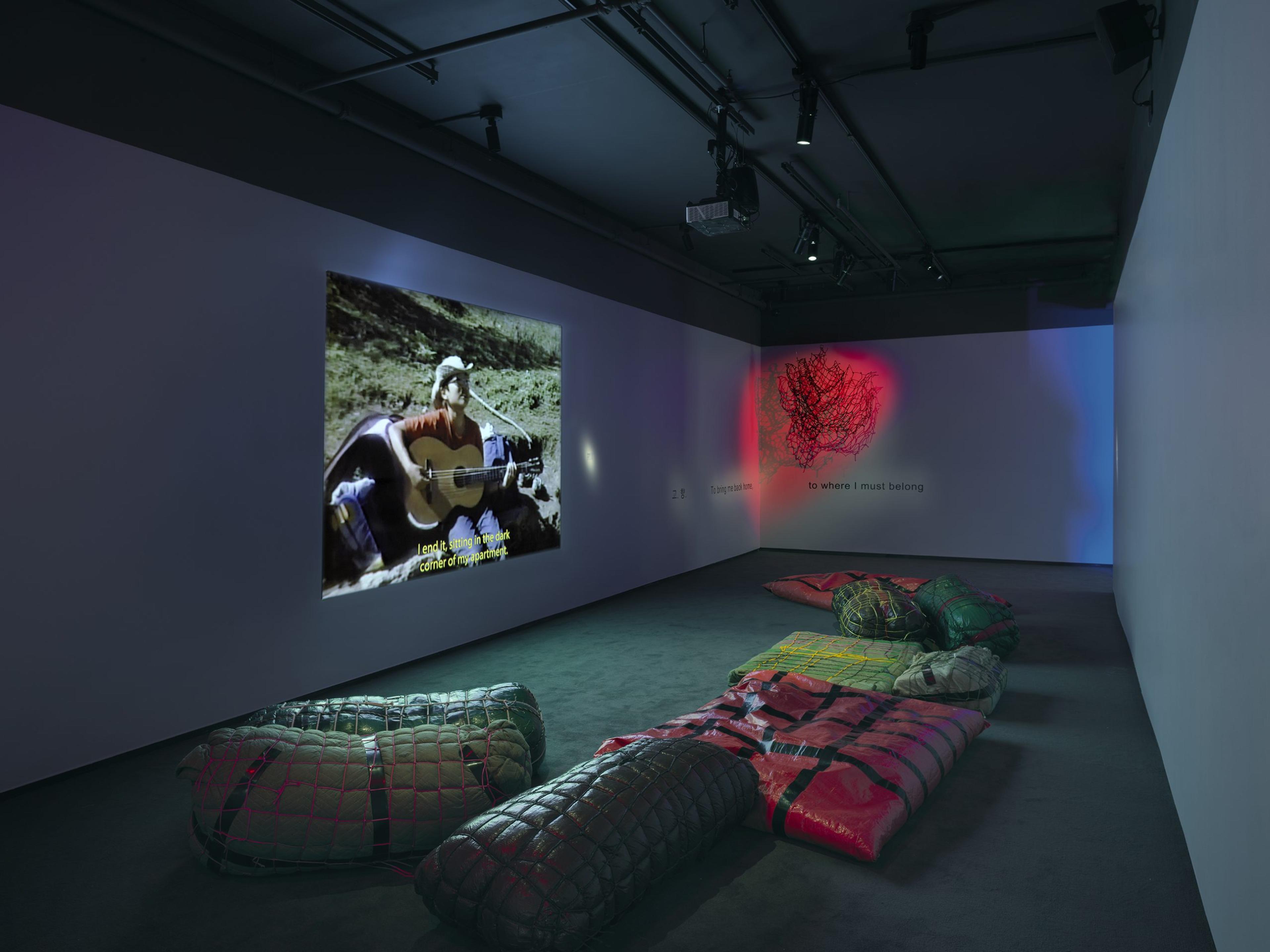 A dark museum gallery room showing a video installation with several sleeping bags on the floor