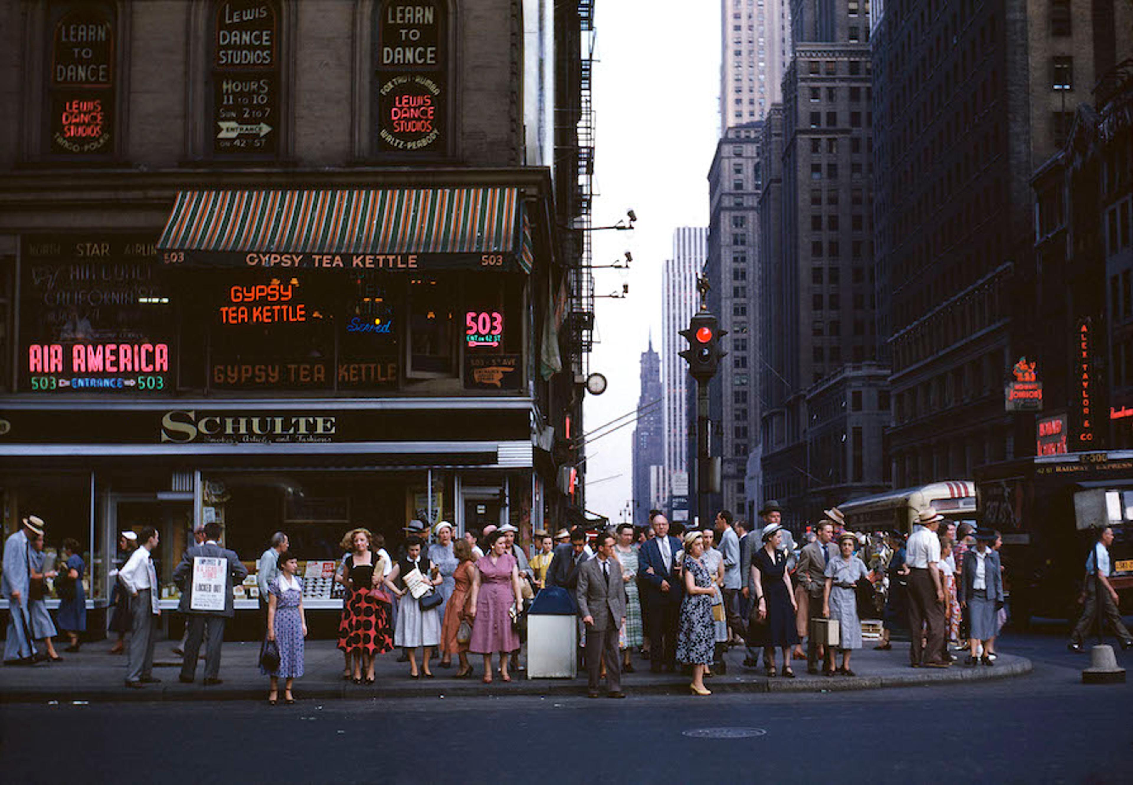 A photograph of a crowd of people standing on a sidewalk on Sixth Avenue in New York City