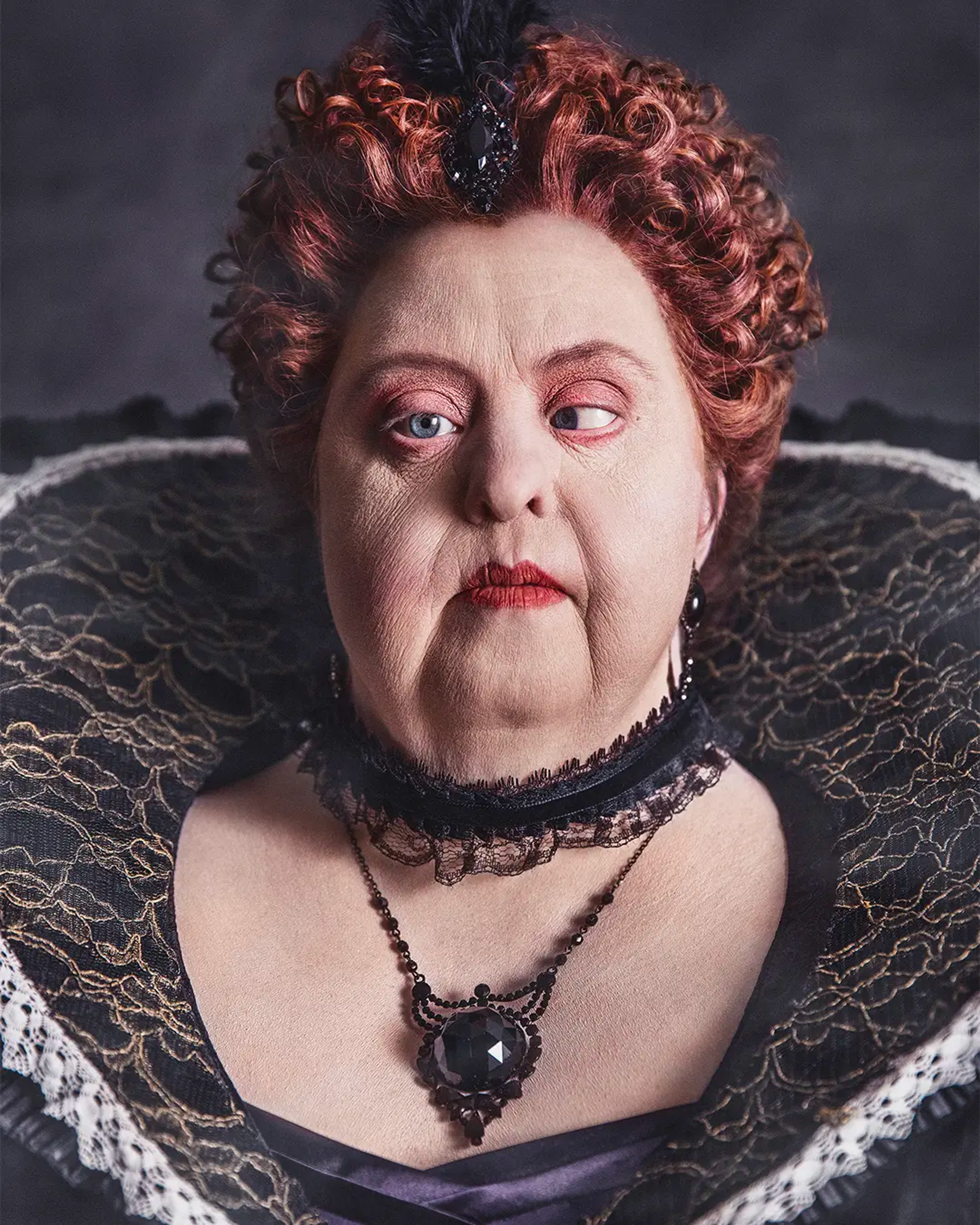 Porträtt av en kvinna i barockstil med uppsatt lockigt hår, dramatisk makeup och svart spetskrage. Hon bär ett stort svart smycke runt halsen.