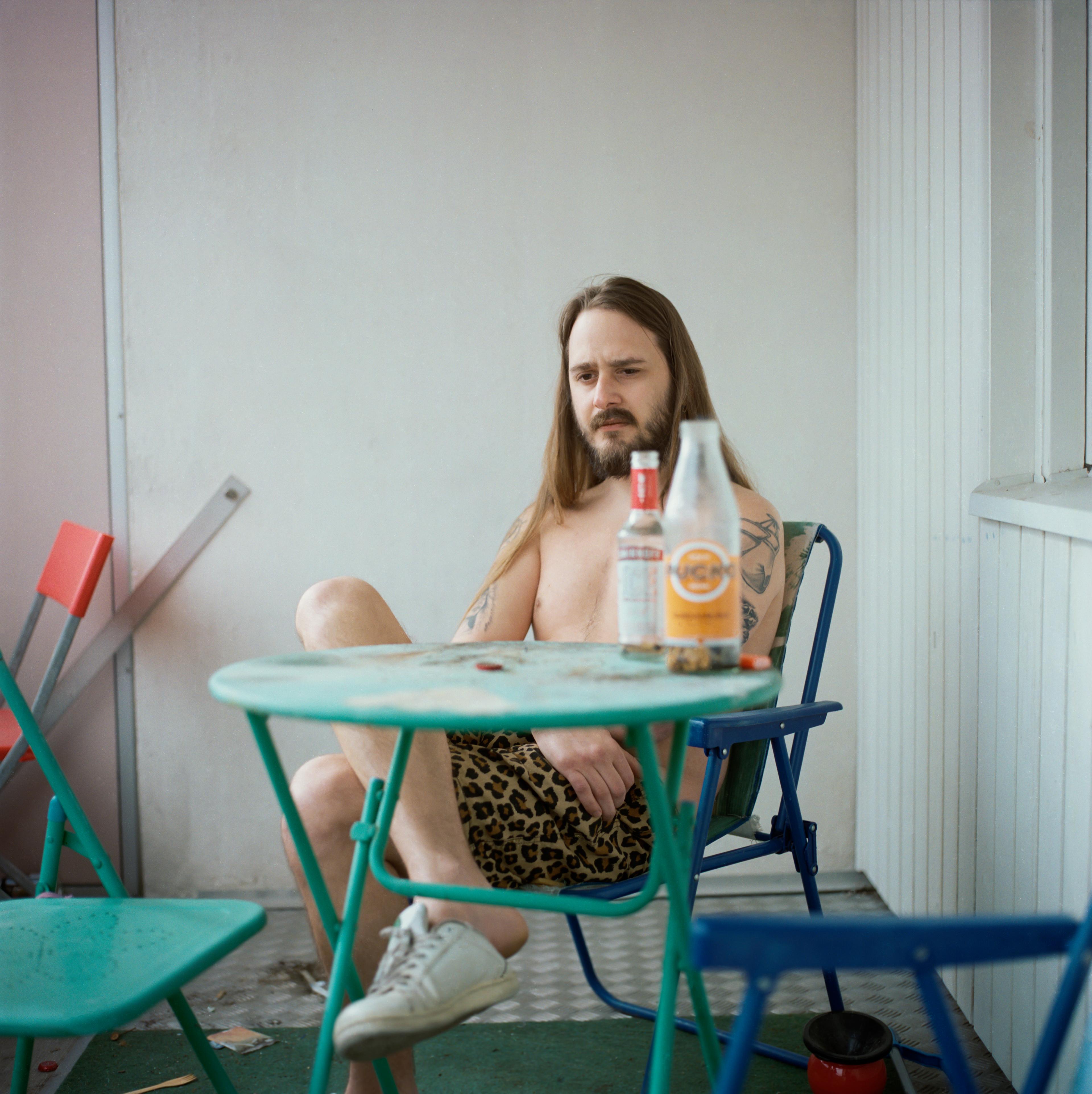 A man with long hair sits on a balcony in a shirt and shorts