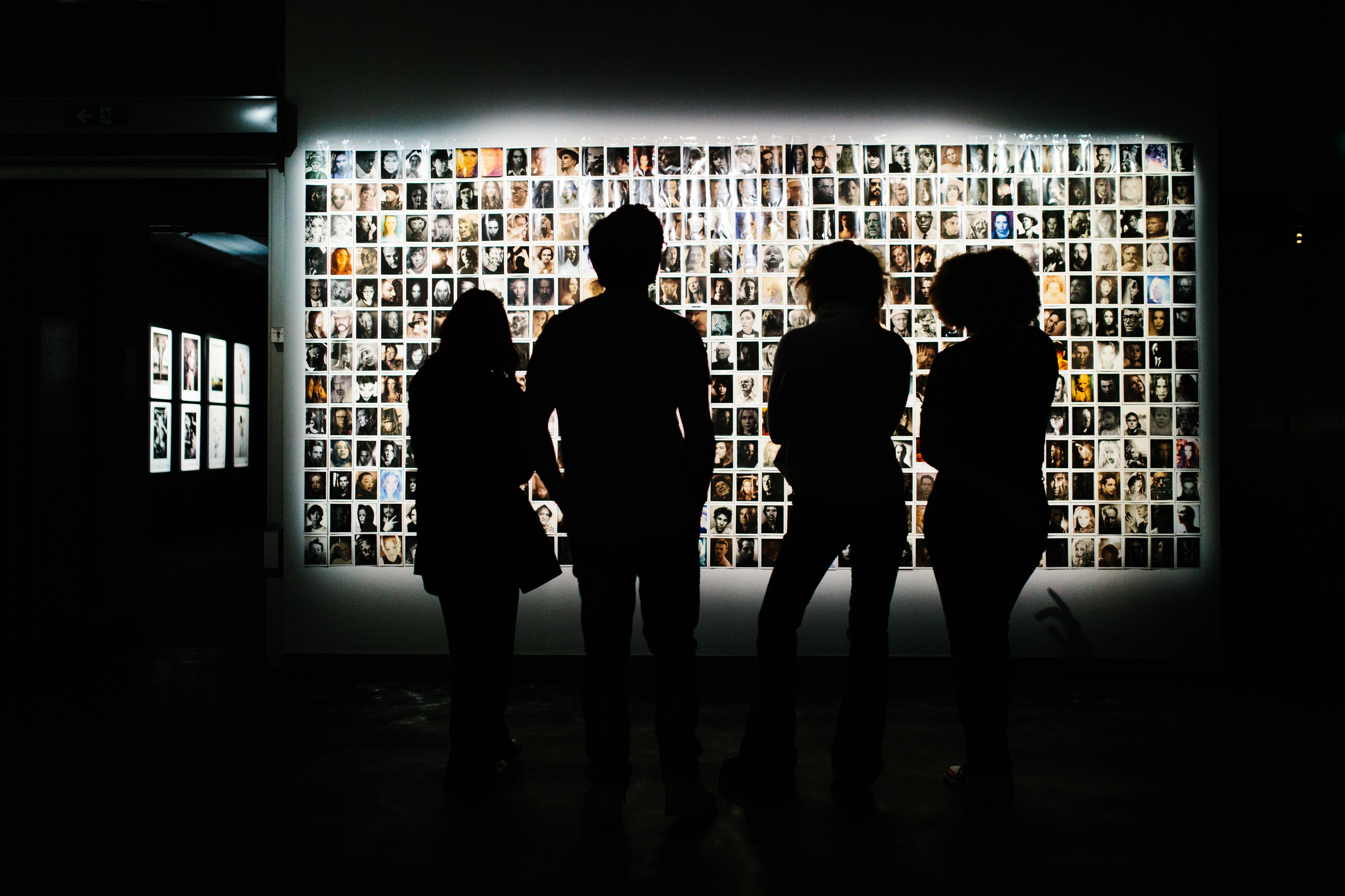 Four people in backlight in front of a collage art piece in a sombre exhibition space 