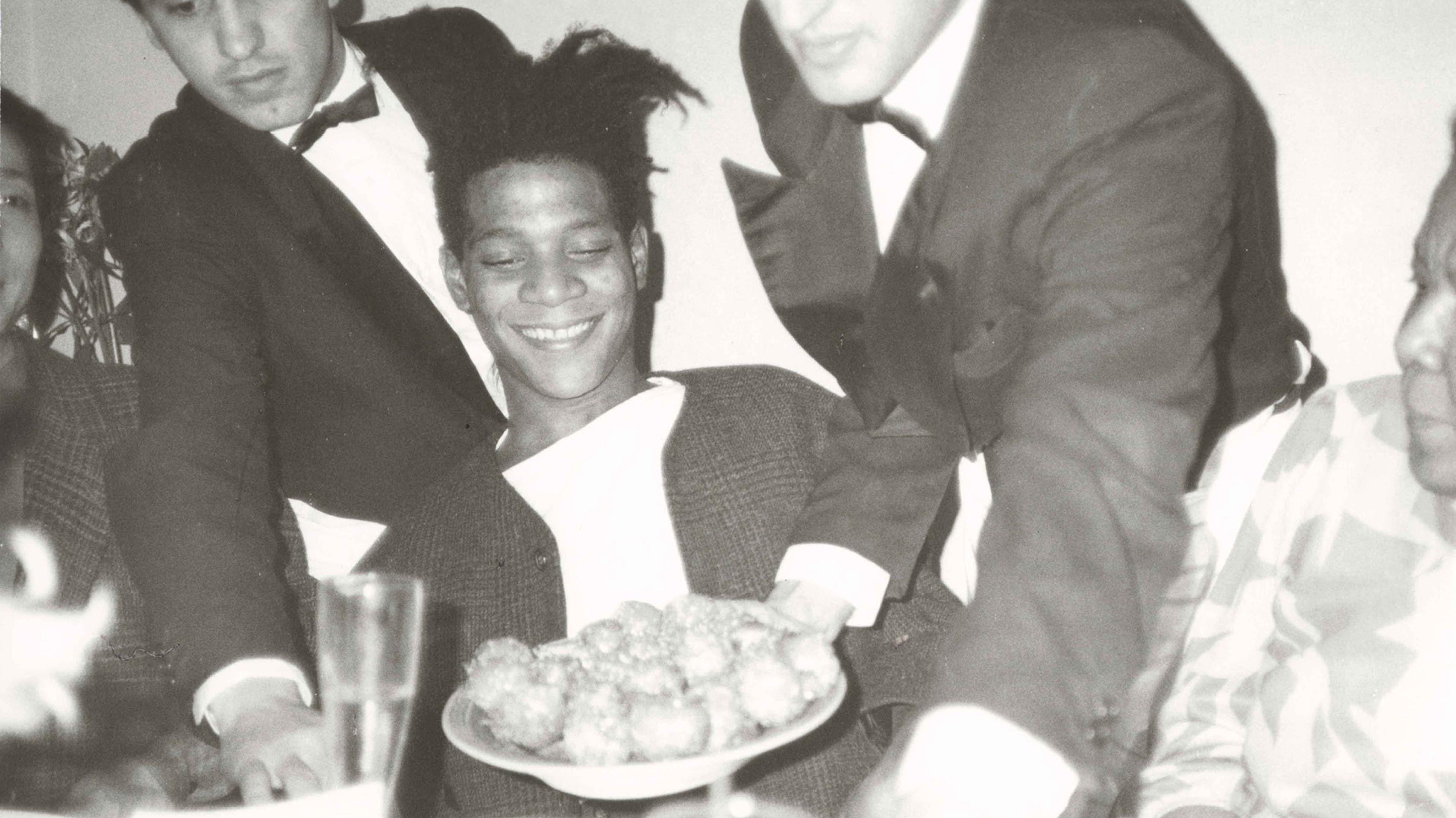 A black-and-white photograph of a smiling young man being handed a plate of food by two waiters in tuxedos