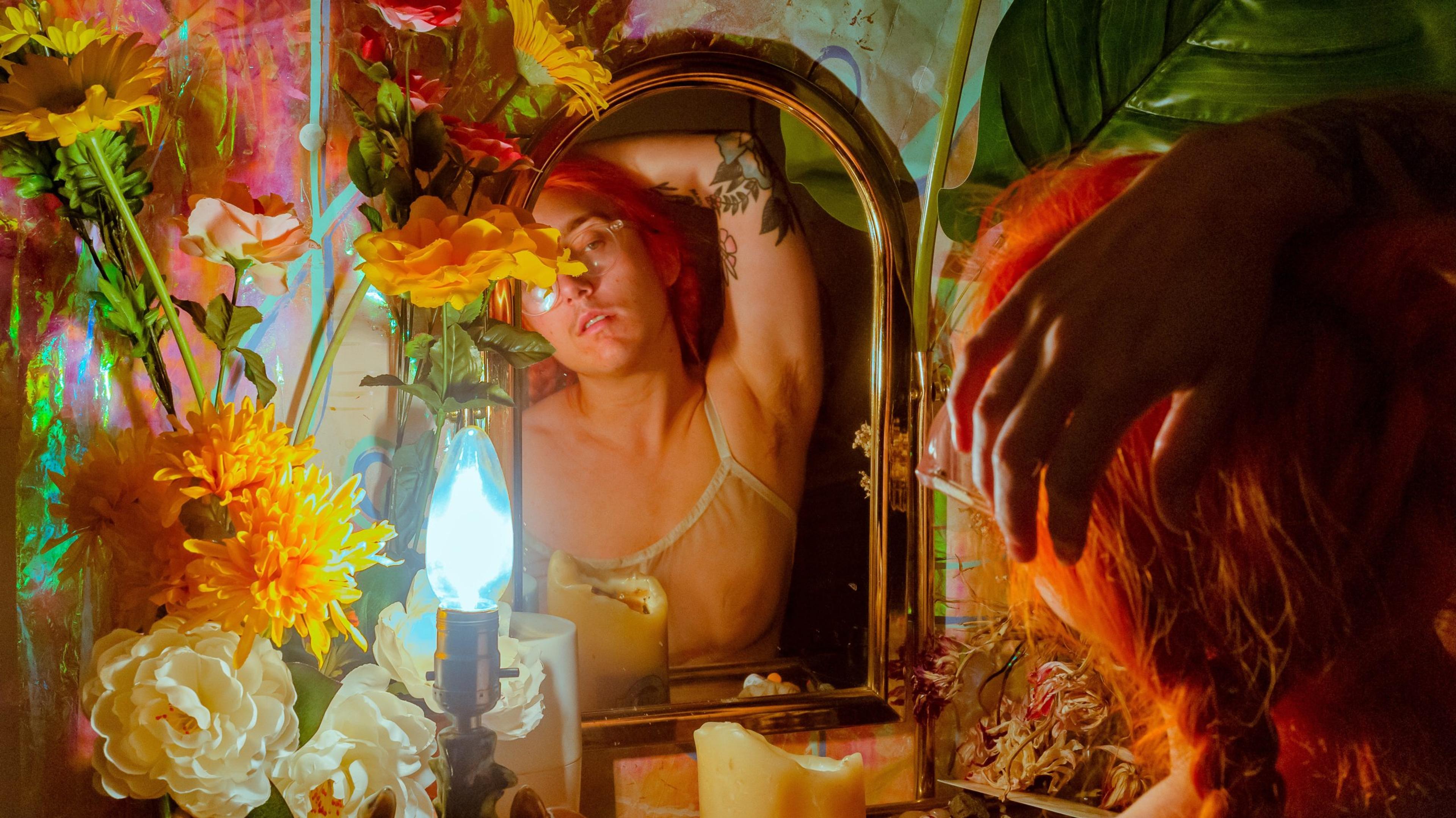 A photograph of a red-headed person looking in a mirror while holding their arm above their head, surrounded by vibrant foliage