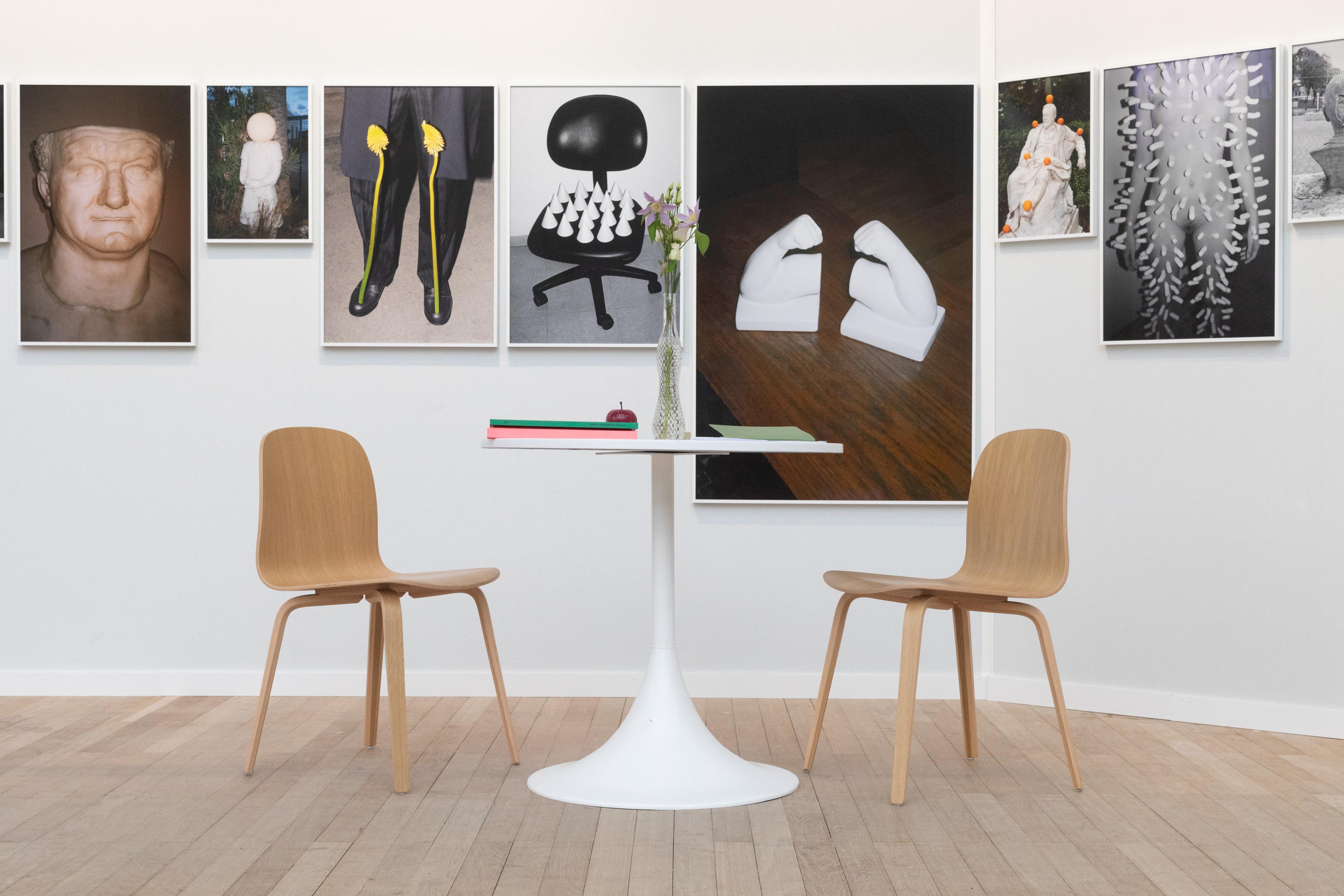 A view of a gallery's exhibiting booth with photographs of varying sizes on the wall behind a white table and light-colored wooden chairs.