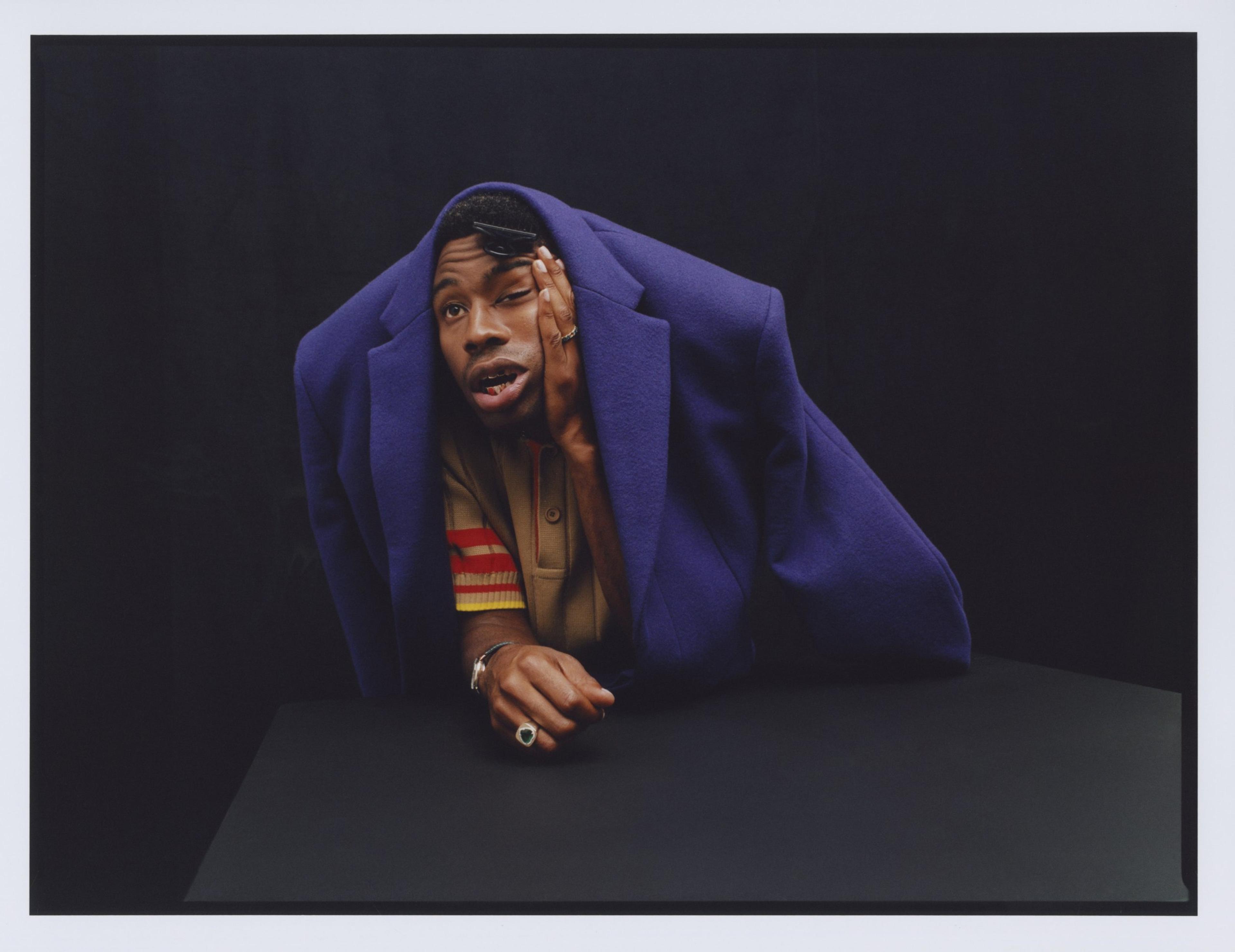 A photograph of Tyler, The Creator with a purple blazer draped on his head and holding his hand to his eye