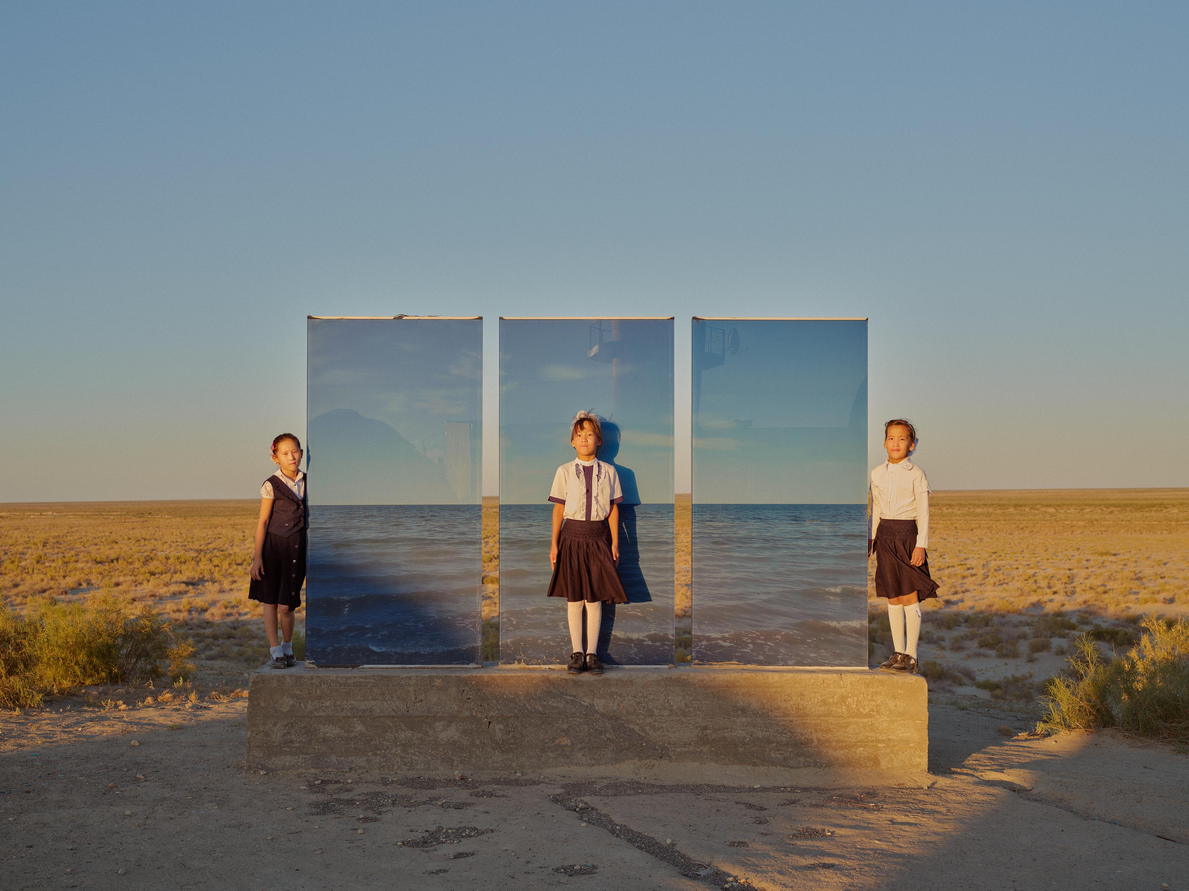 Three young girls in three different styles of school uniforms standing next to and in front of three screens of artwork depicting the sea and horizons. The girls and artworks are standing on on concrete block which is placed on a prairie like landscape.