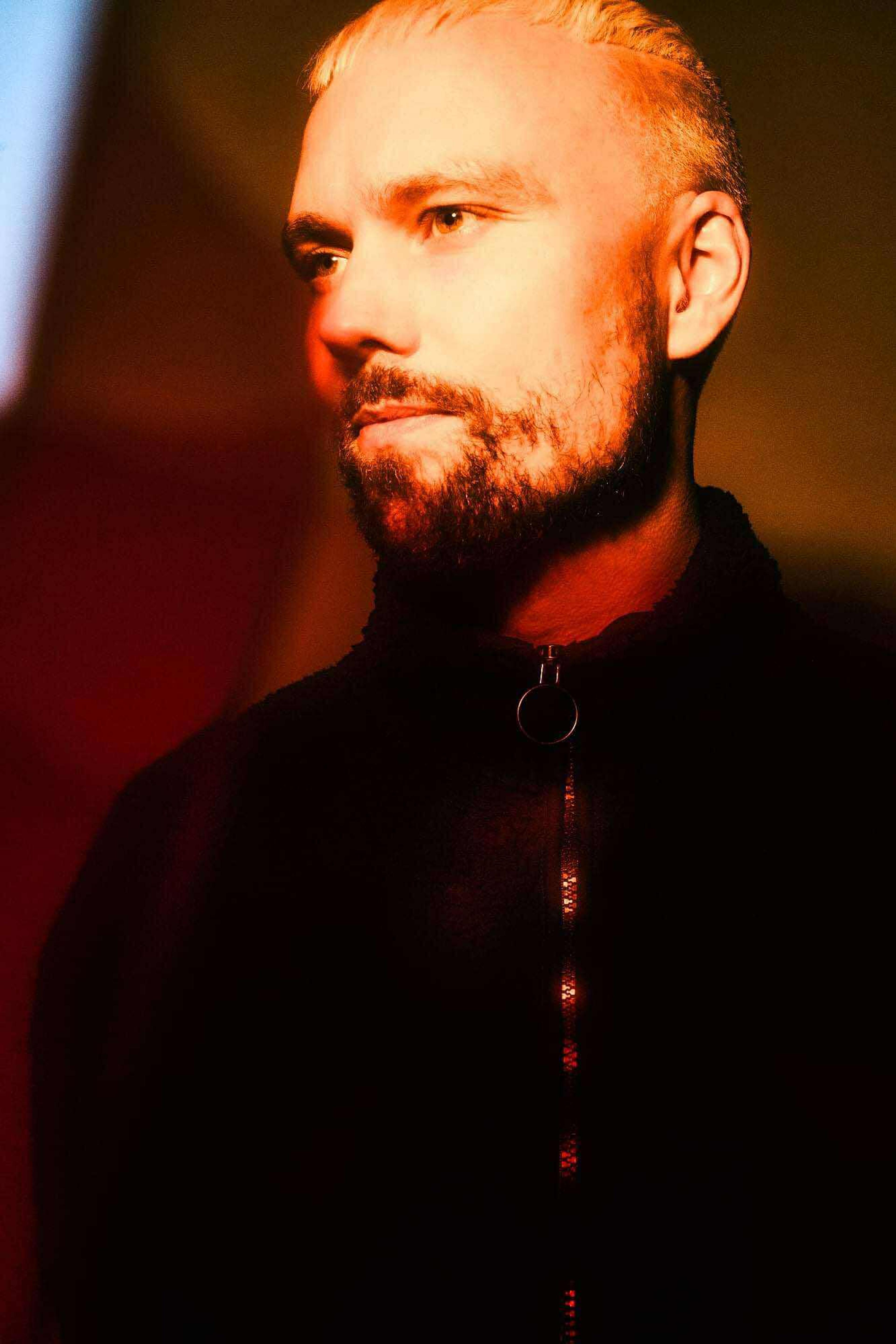 Portrait of Eyvind Blix under warm, dramatic lighting, wearing a dark zip-up top with a circular zipper pull