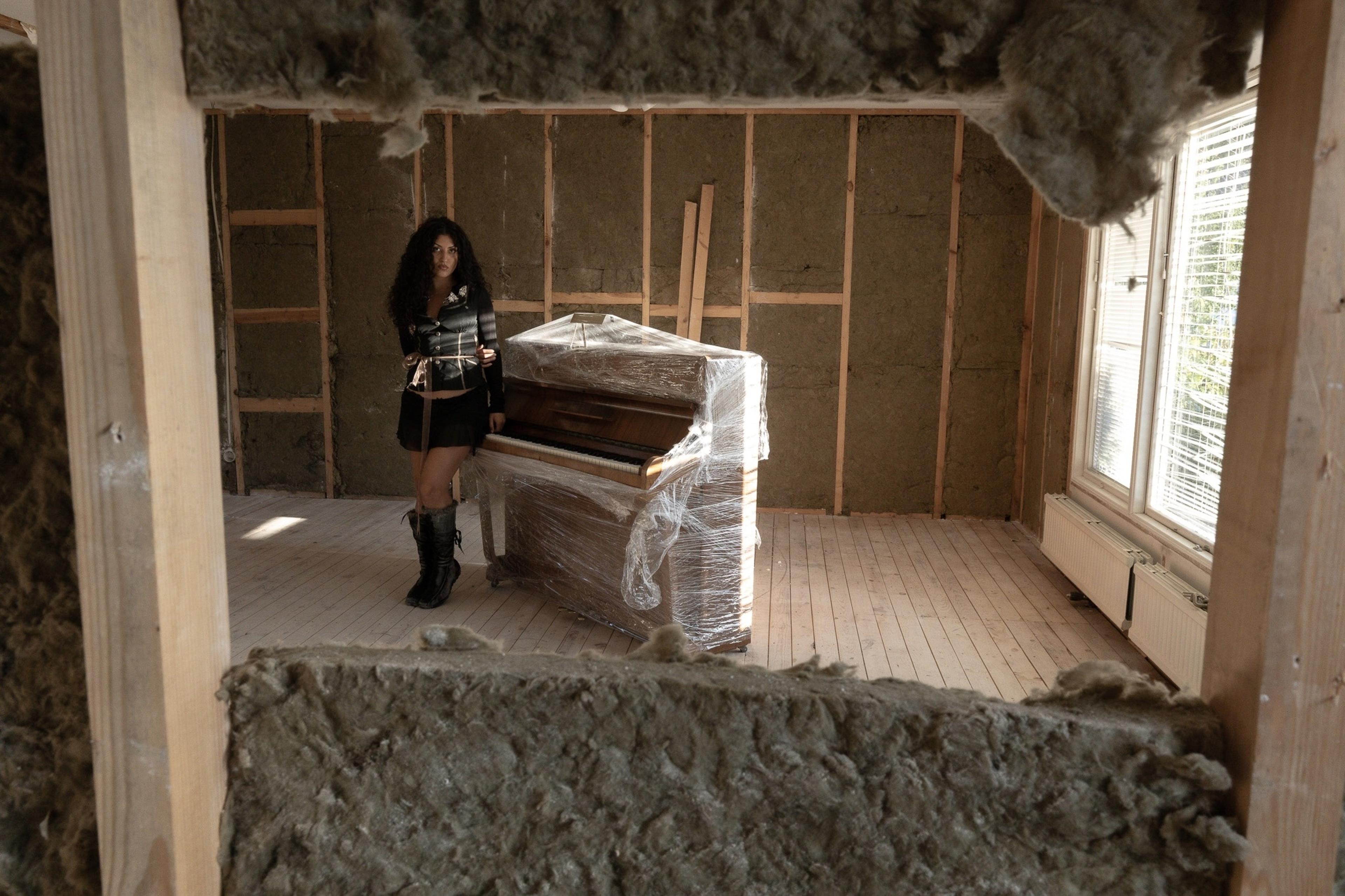 Venus Anon in a room stripped of its walls, leaning against a piano wrapped in plastic.