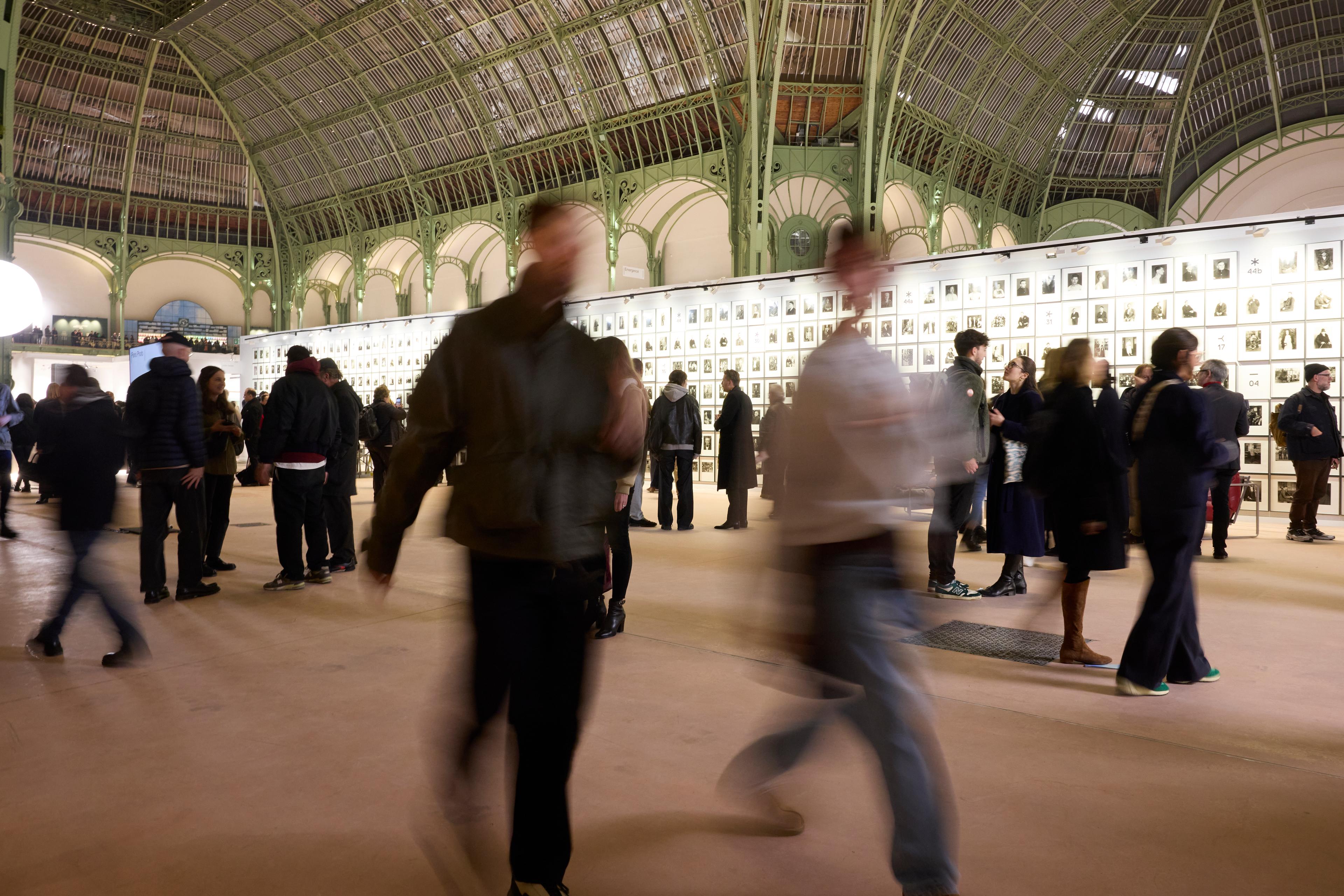 A motion-blurred view of people walking in front of a gallery wall featuring hundreds of small framed photographs