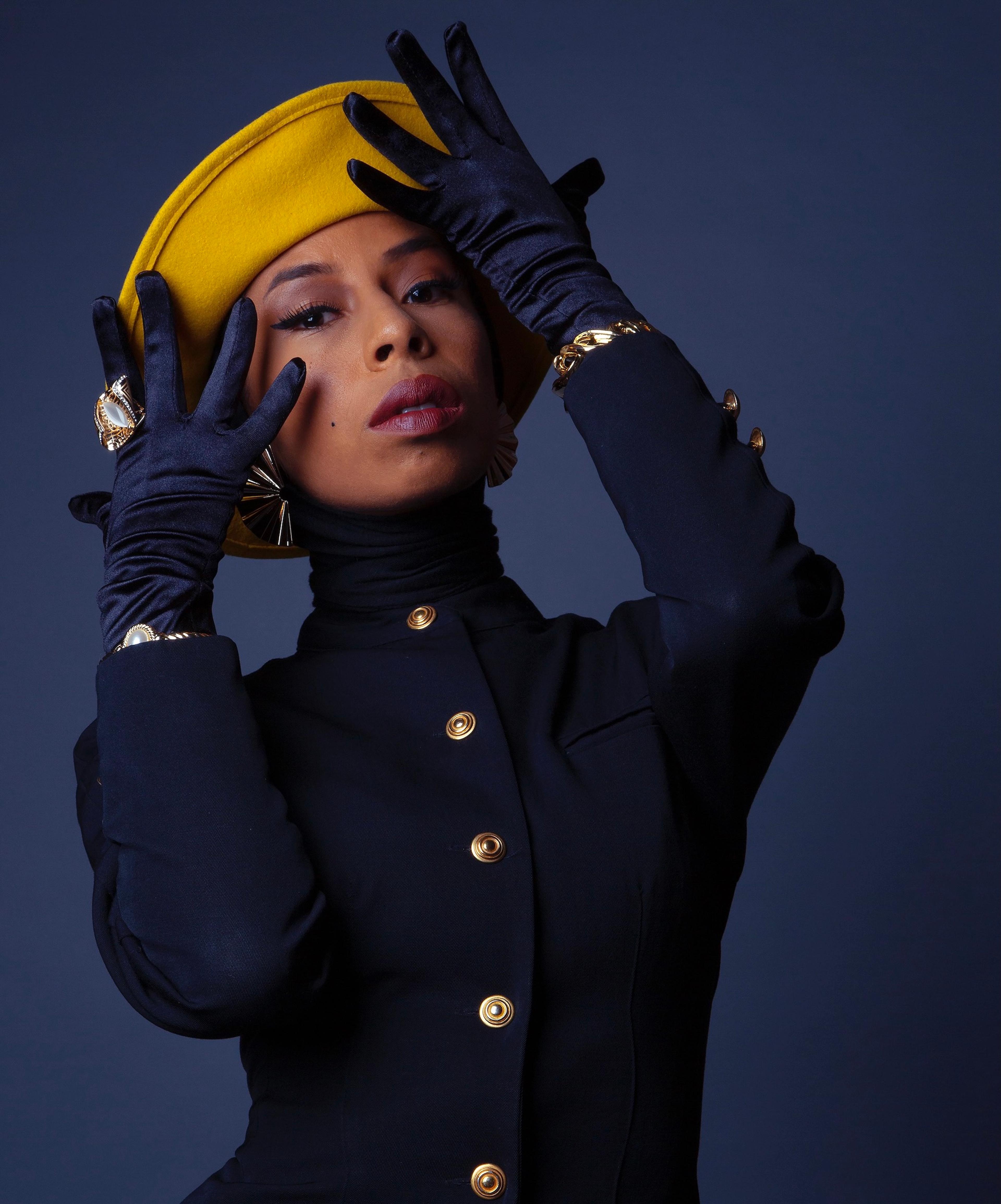 A portrait of a woman wearing a suit, gloves, and yellow cap against a navy blue background