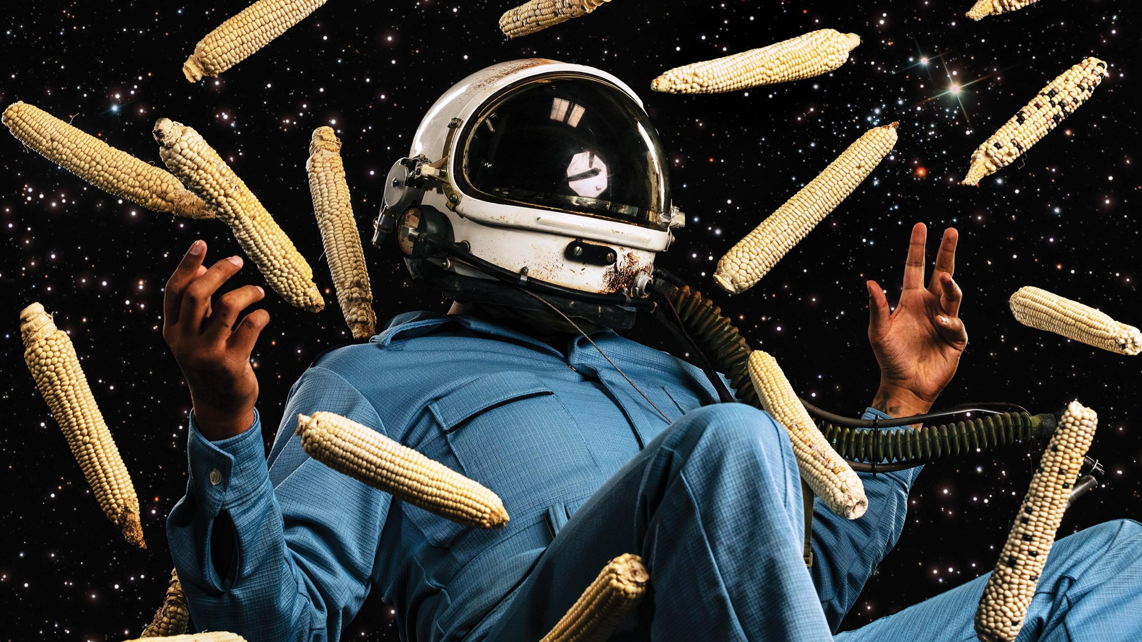 A photographic artwork of a man in a blue space suit and helmet is suspended in the air, surrounded by floating corn cobs