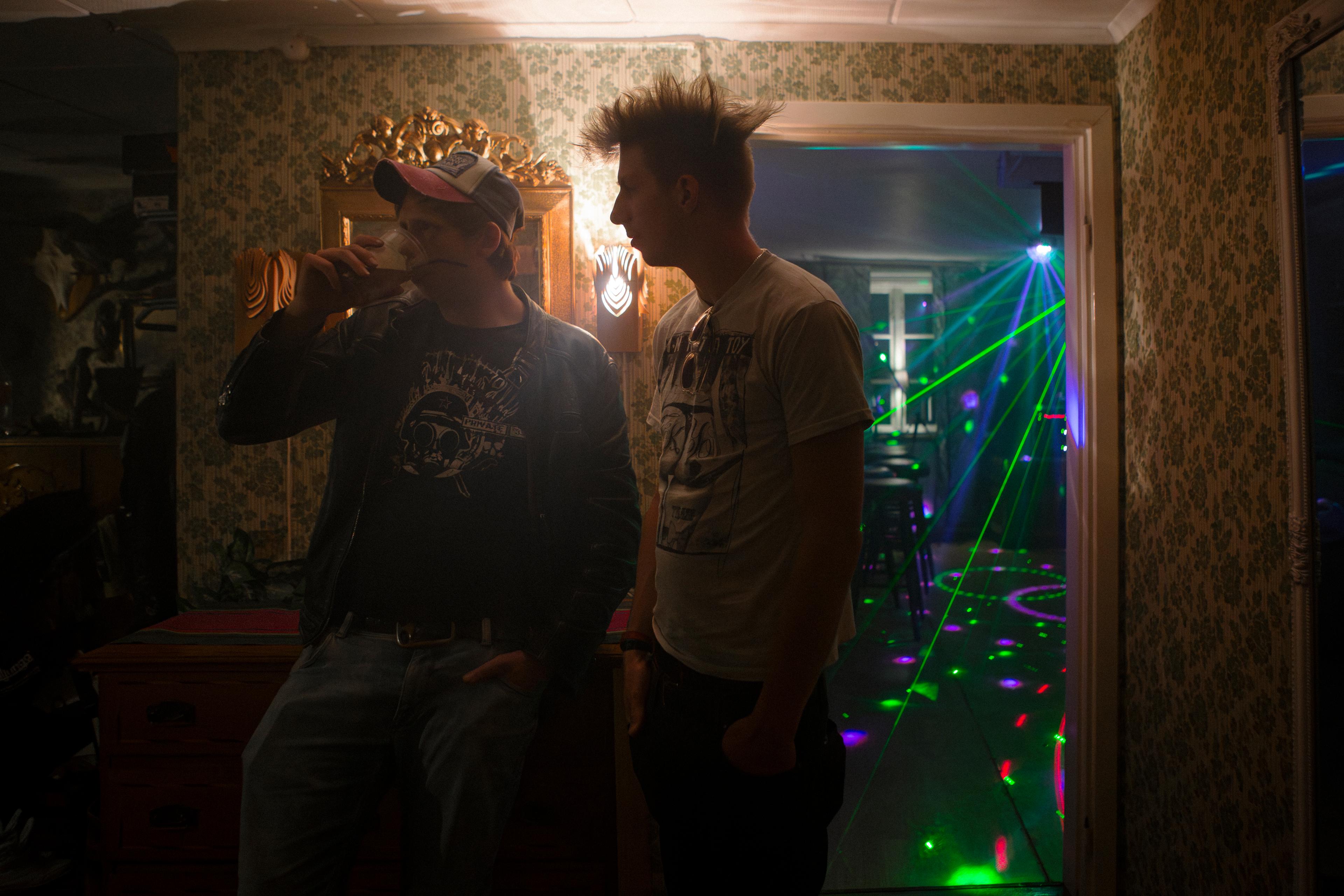 Two people are standing in a living room, it seems to be a house party, some neon lights in the background. one guy is wearing a mohawk and tshirt, the other guy is wearing a cap and is drinking beer 