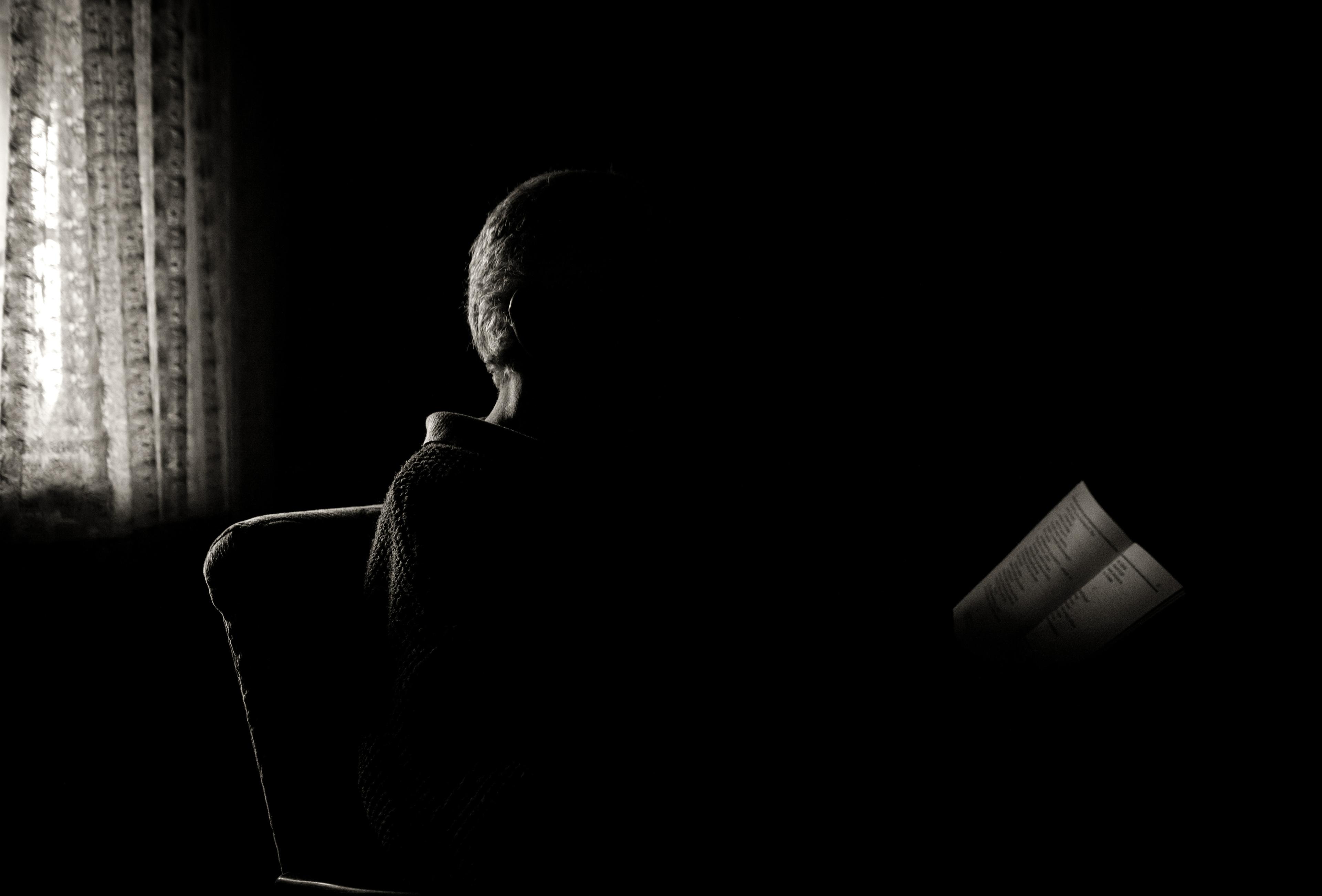 Silhouette of the back of a man captured in a dark, almost black room with only light coming from a curtain