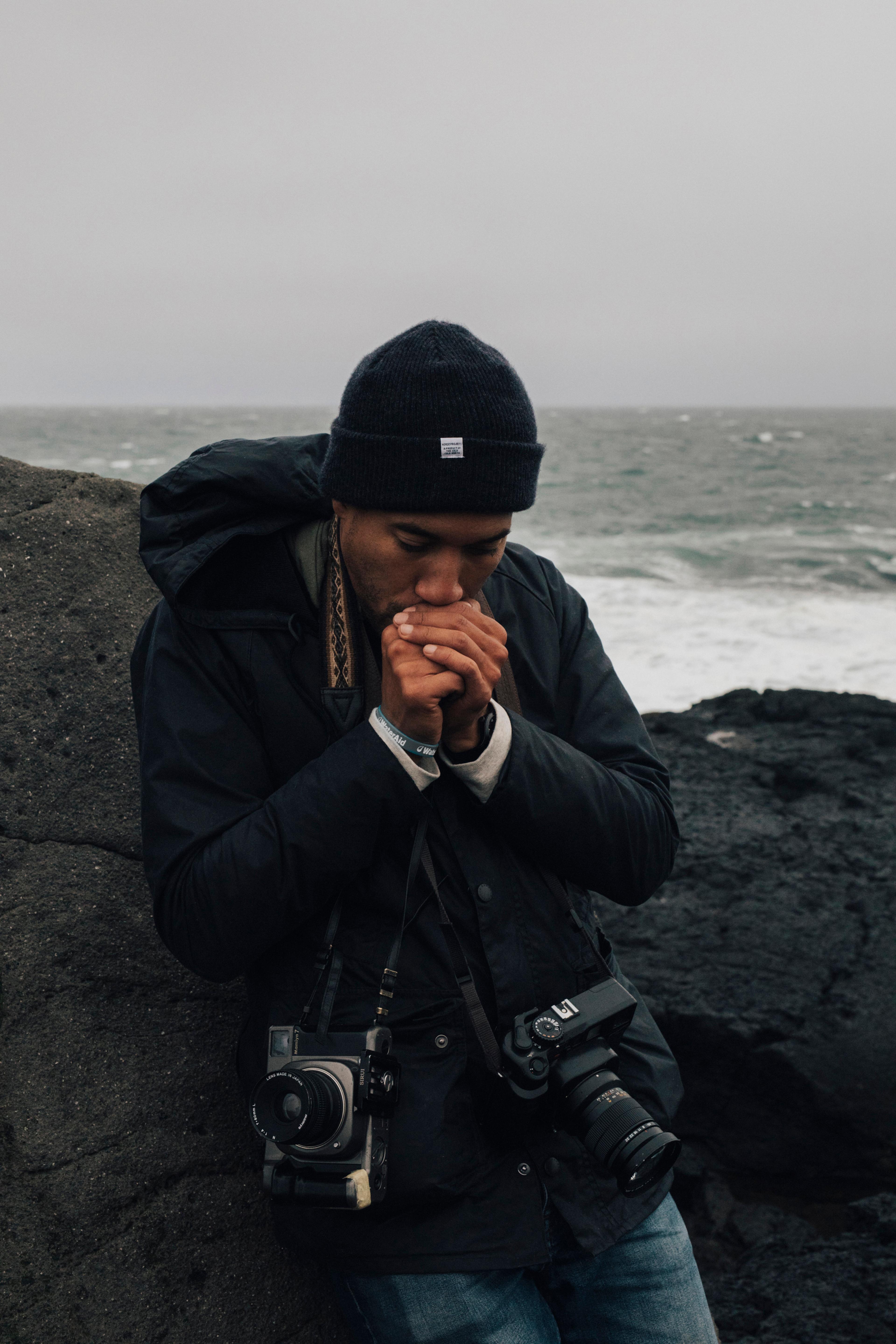 Photo of Mustafah Abdulaziz leaning on black rocks in a dark outfit with the sea in the background