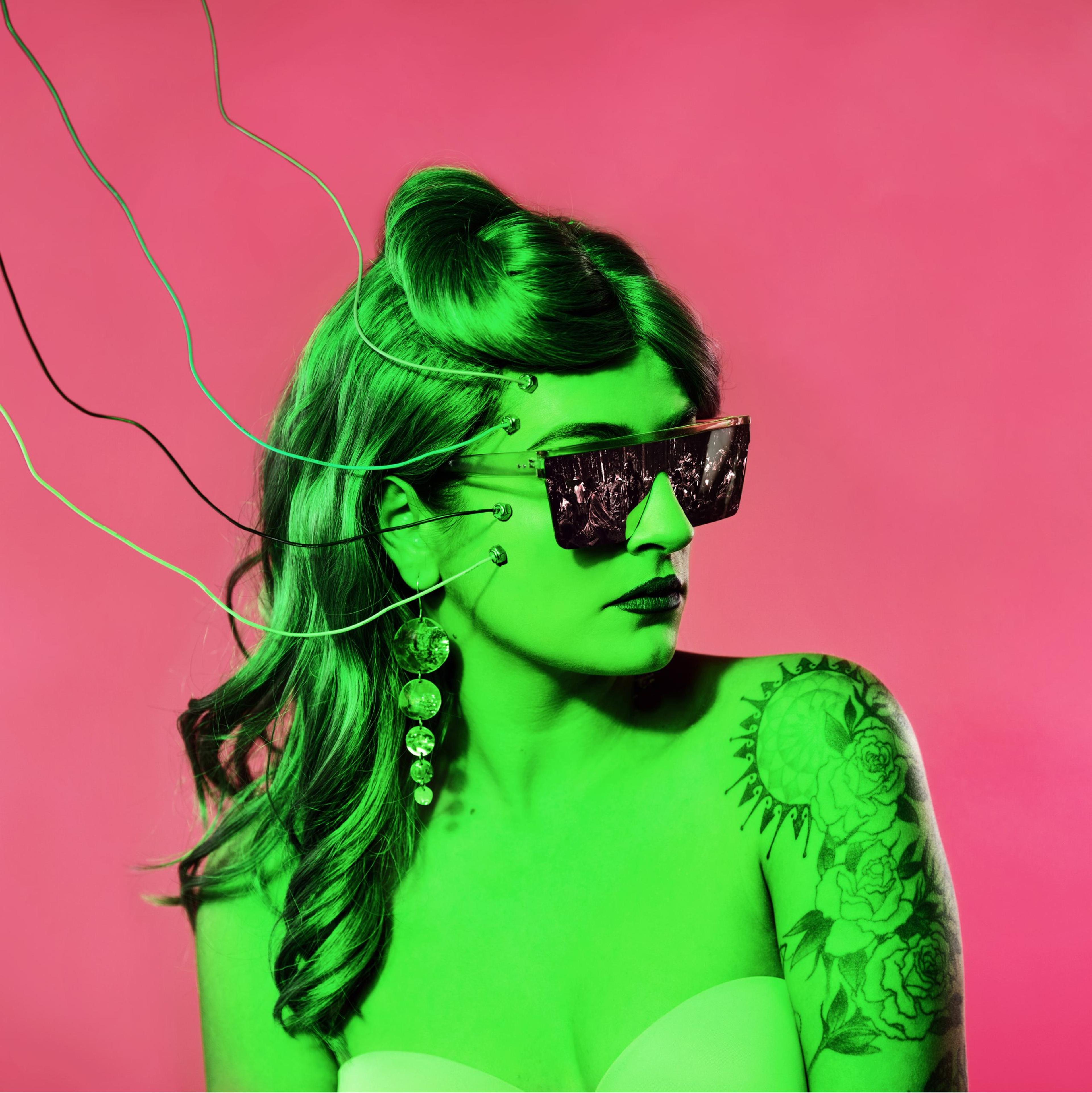 A photograph of a woman against a pink background, wearing sunglasses and illuminated by green lighting with electrodes attached to her face