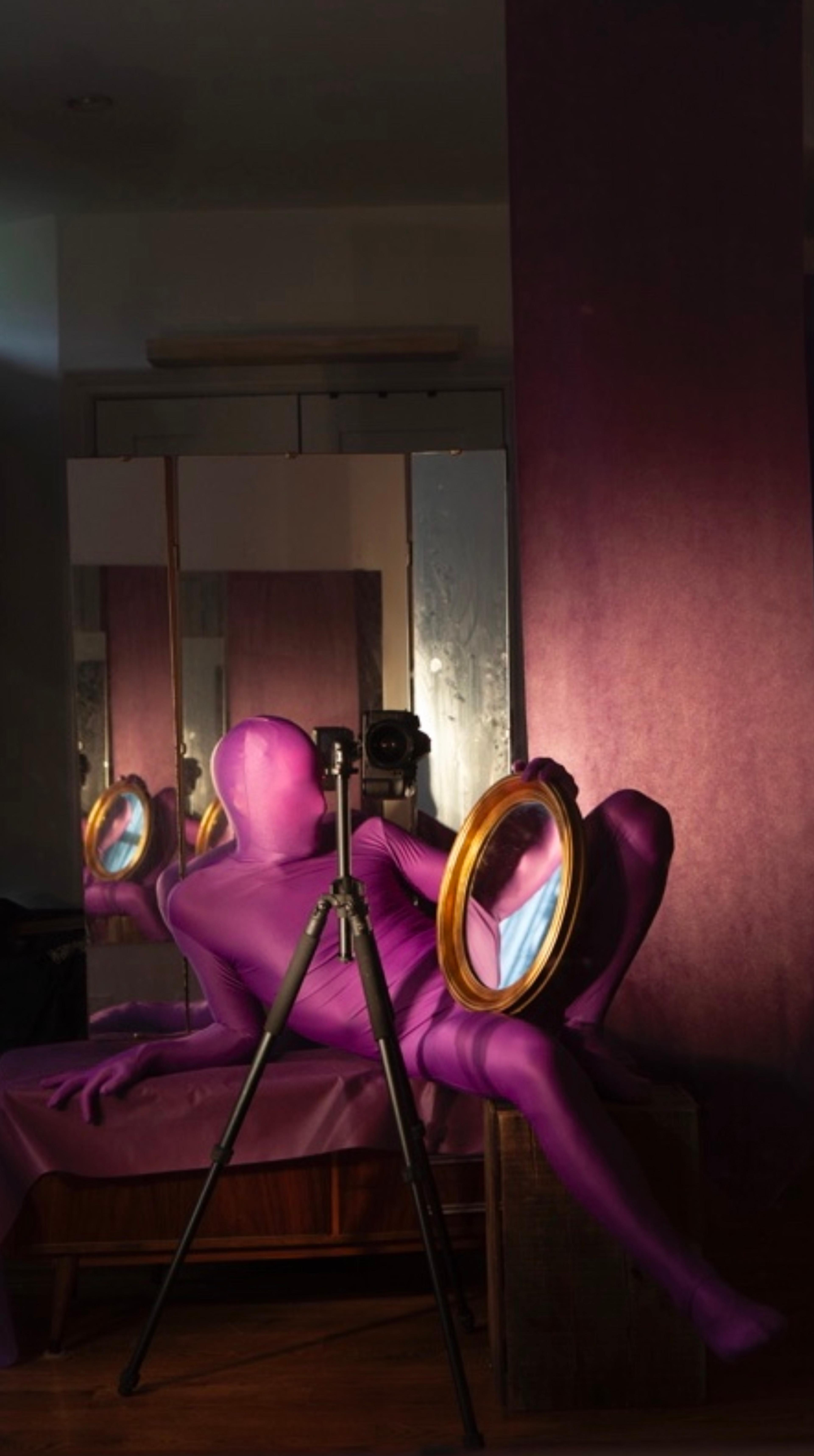 A photograph of a person in a pink full-body suit lounges in front of a camera and a mirror